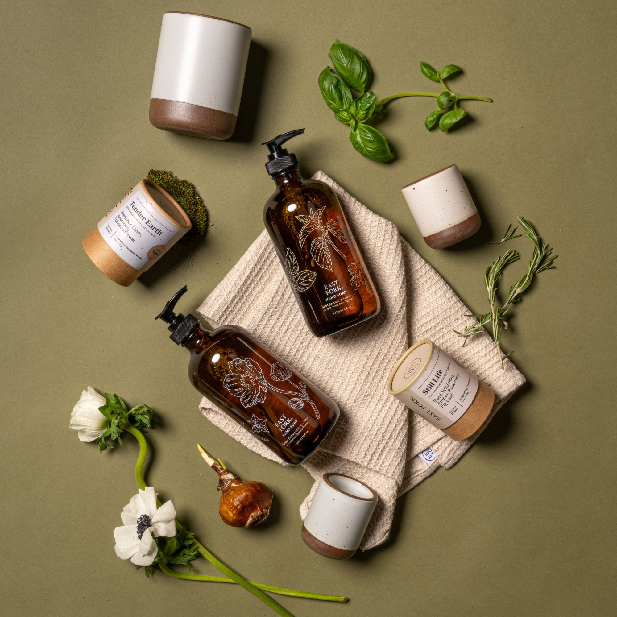 Two amber glass soap bottles with candles, towels, and herbal accents arranged on a green surface