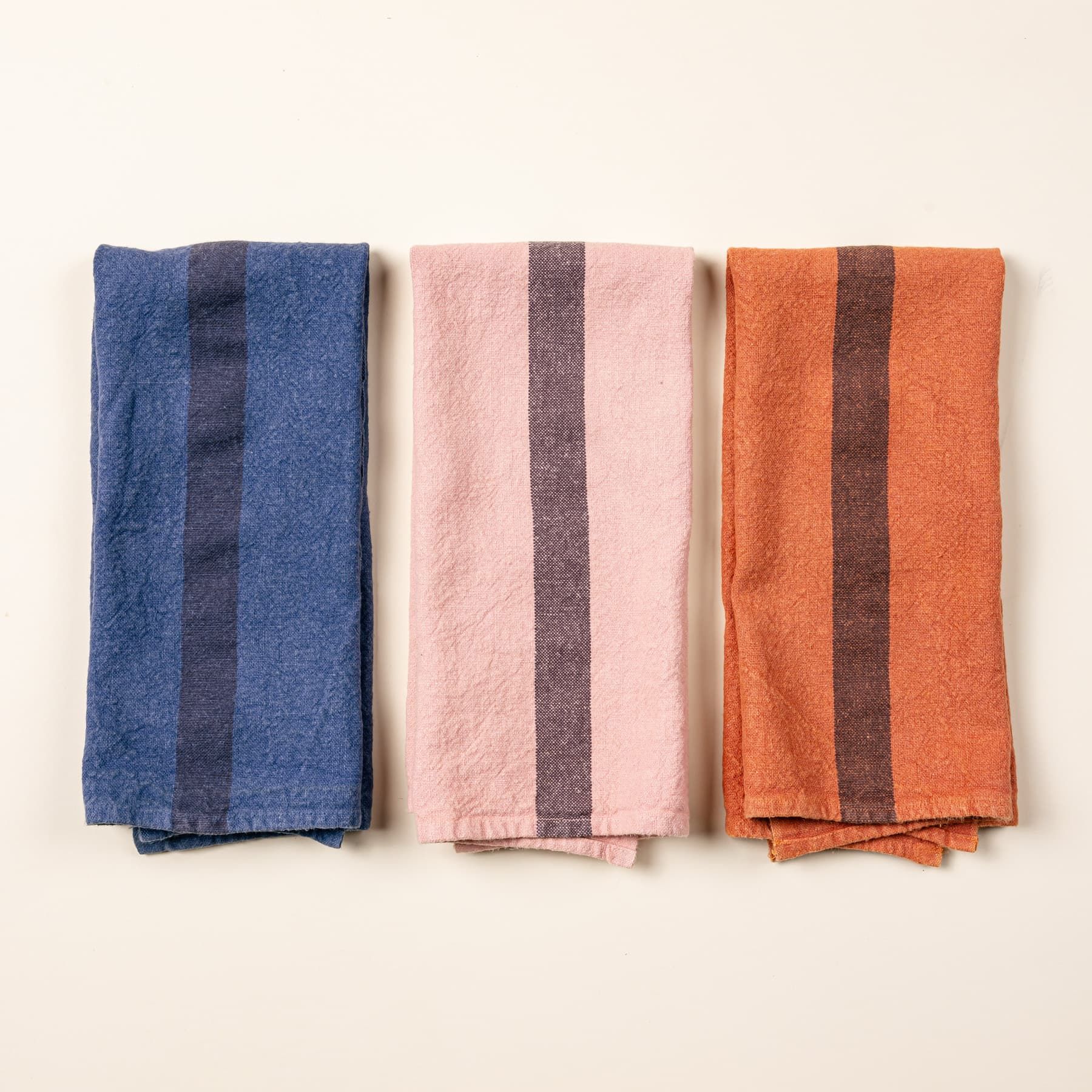 3 folded linen towels in an indigo, salmon, and terracotta color with a plum stripe in the middle of each