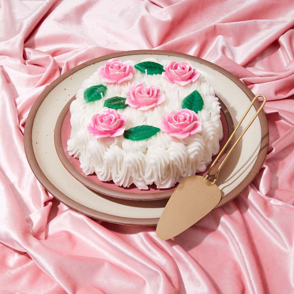 A shiny pink cloth is draped under plates topped by a candle that has the size, shape and color of a cake with icing, all pink and white with green stems for the wax roses
