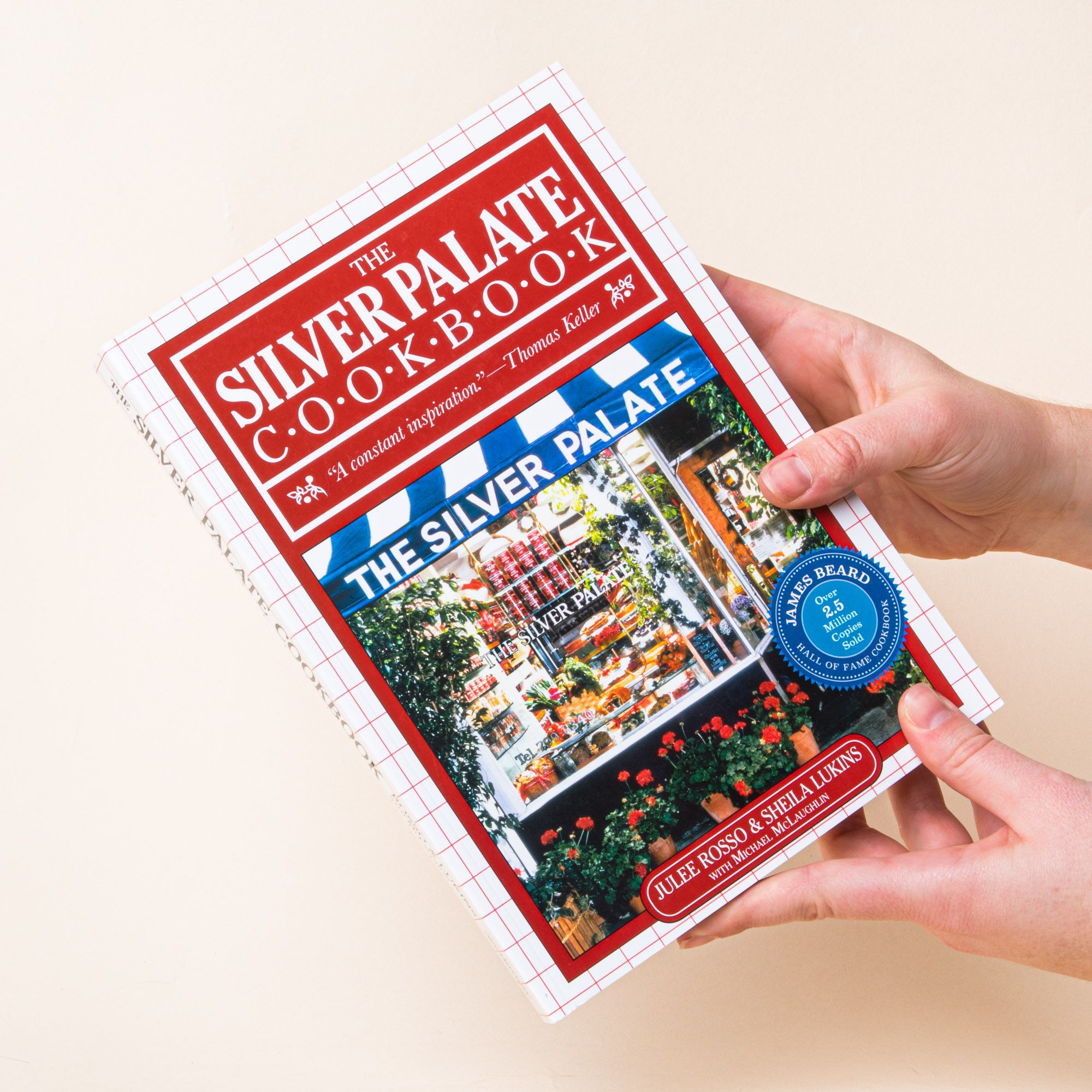Hands holding a book with a red cover and a photo of a storefront and outside garden, the title reads "The Silver Palate Cookbook"