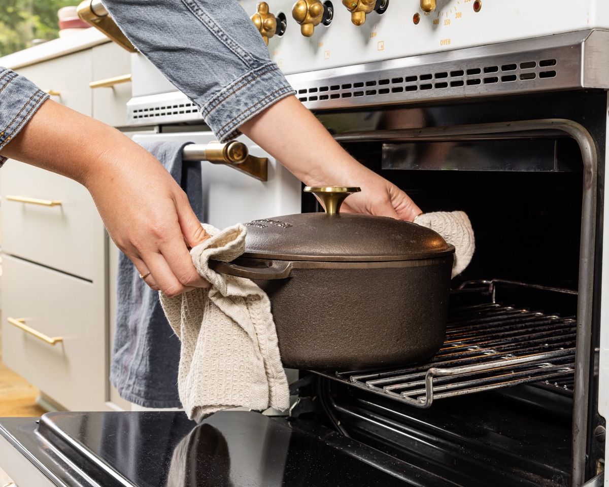 Hands hold a kitchen towel to carry a cast iron dutch oven with a handle on each side and cast iron lid out of an oven.