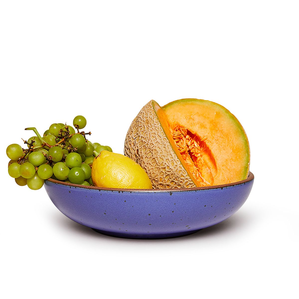 A sliced cantaloupe, whole lemon, and green grapes in a large shallow serving ceramic bowl in a true cool blue color featuring iron speckles and an unglazed rim.