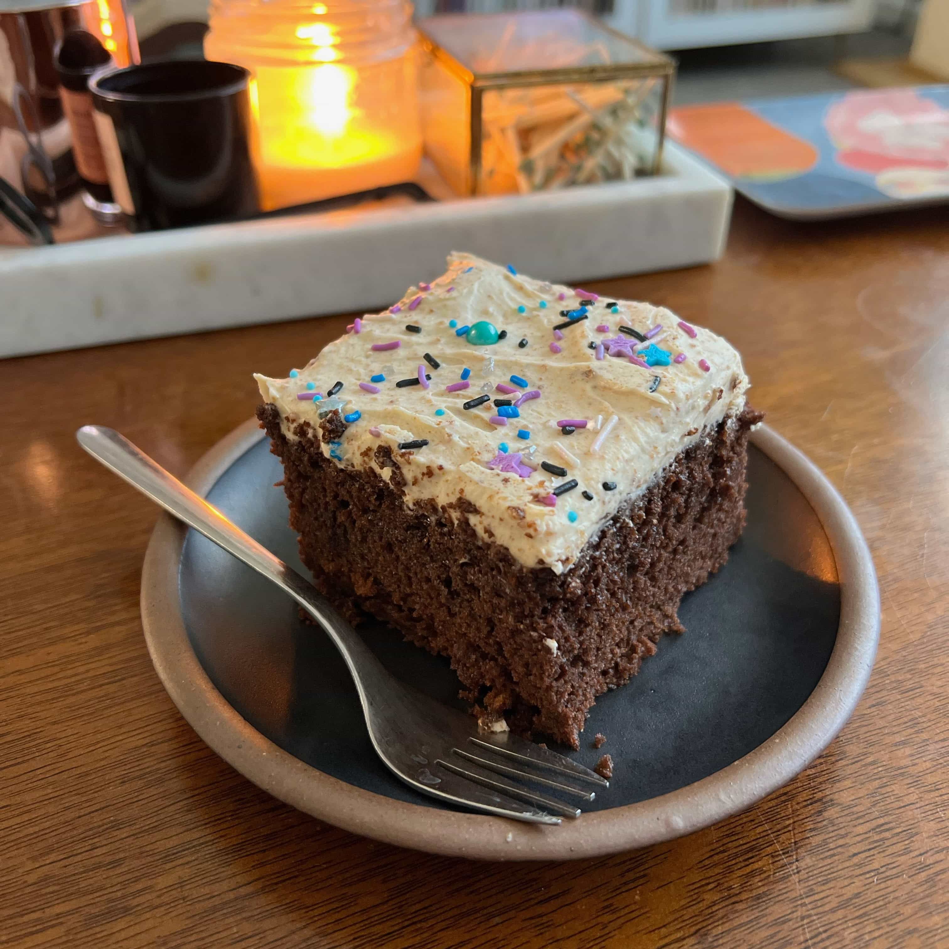 A slice of chocolate cake with cream frosting and sprinkles sits on a black ceramic cake plate with a fork.