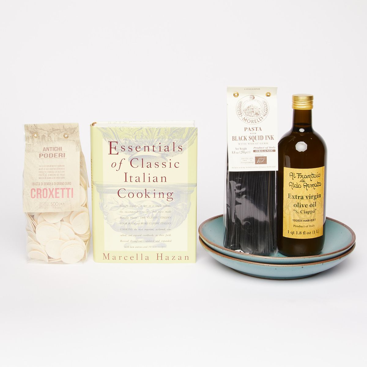 From left to right: A clear bag of round-shaped pasta, a book cover that reads "Essentials of Classic Italian Cooking", two stacked turquoise coupes holding a bag of black squid ink linguine and a dark bottle of olive oil