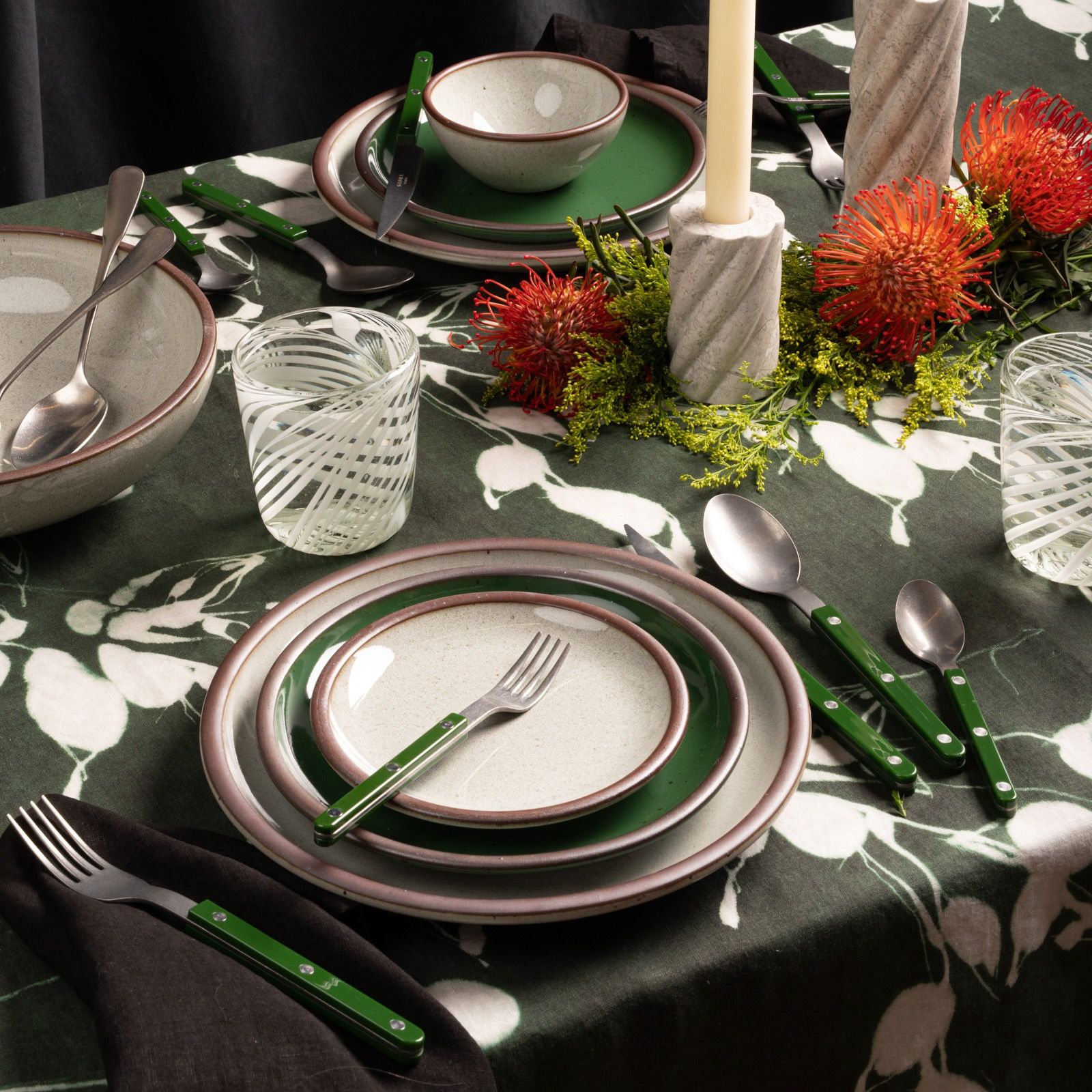 Festive table with icy white and green East Fork pottery, floral accents, marble candles, and green flatware on a dark botanical cloth.