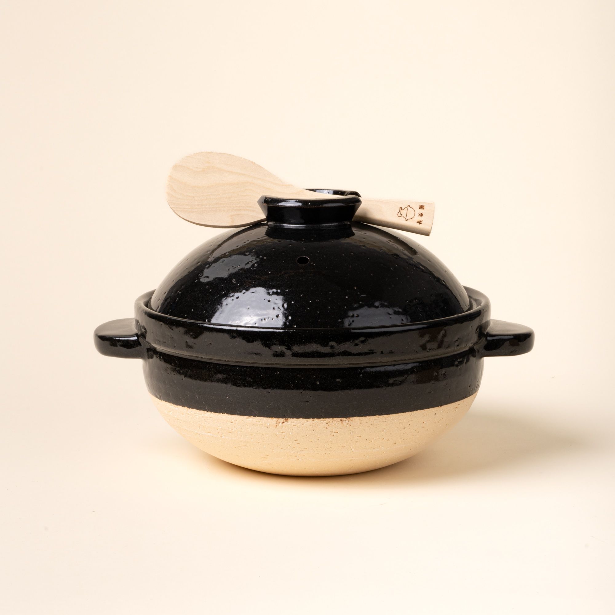 A black-and-neutral color claypot with handles and a shallow-dome shaped lid. The cooking vessel sits atop a wooden board next to a wooden spoon.