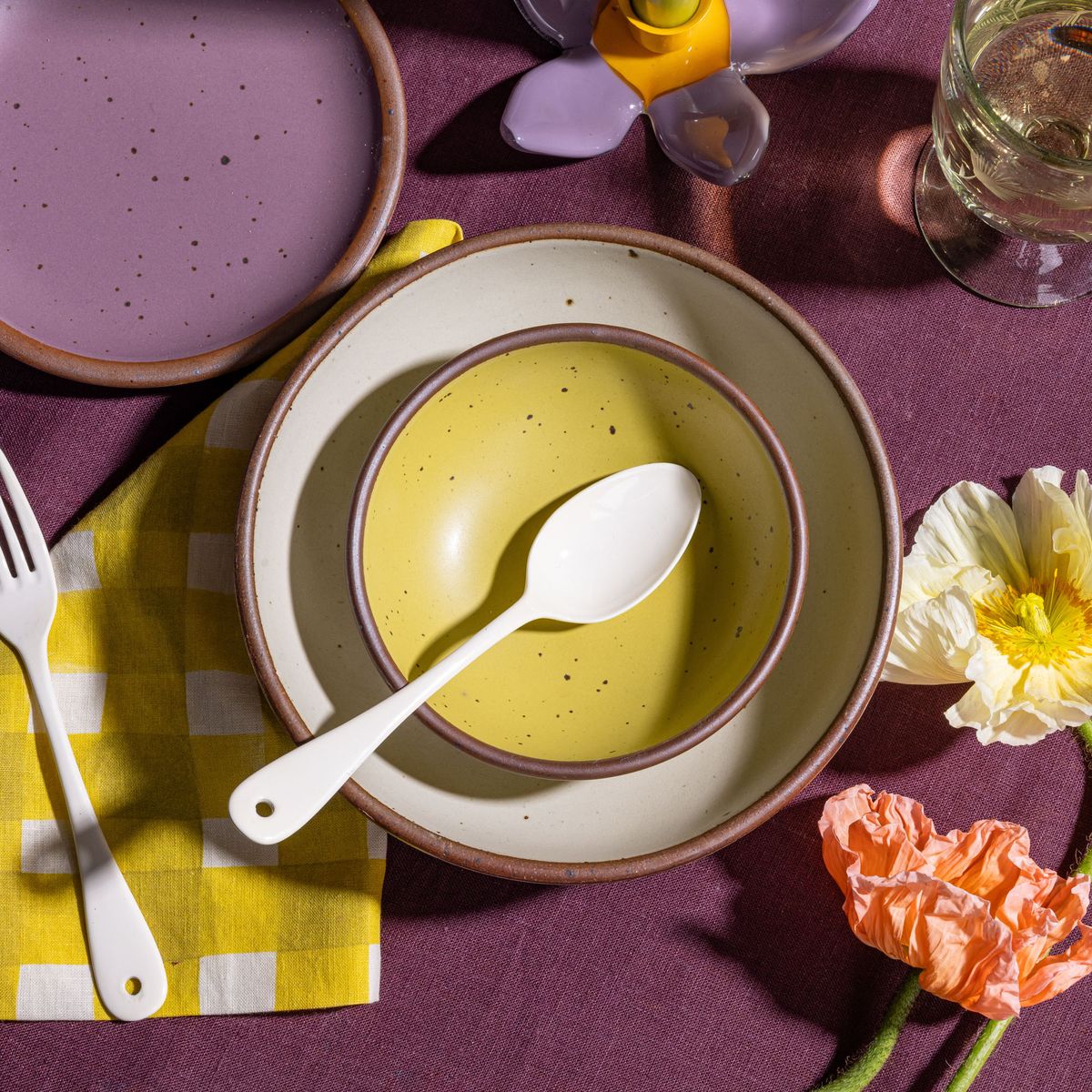 A small dessert sized rounded ceramic bowl in a zesty yellow color nestled in a larger cream ceramic bowl with an enamel spoon, and yellow and purple table linens.