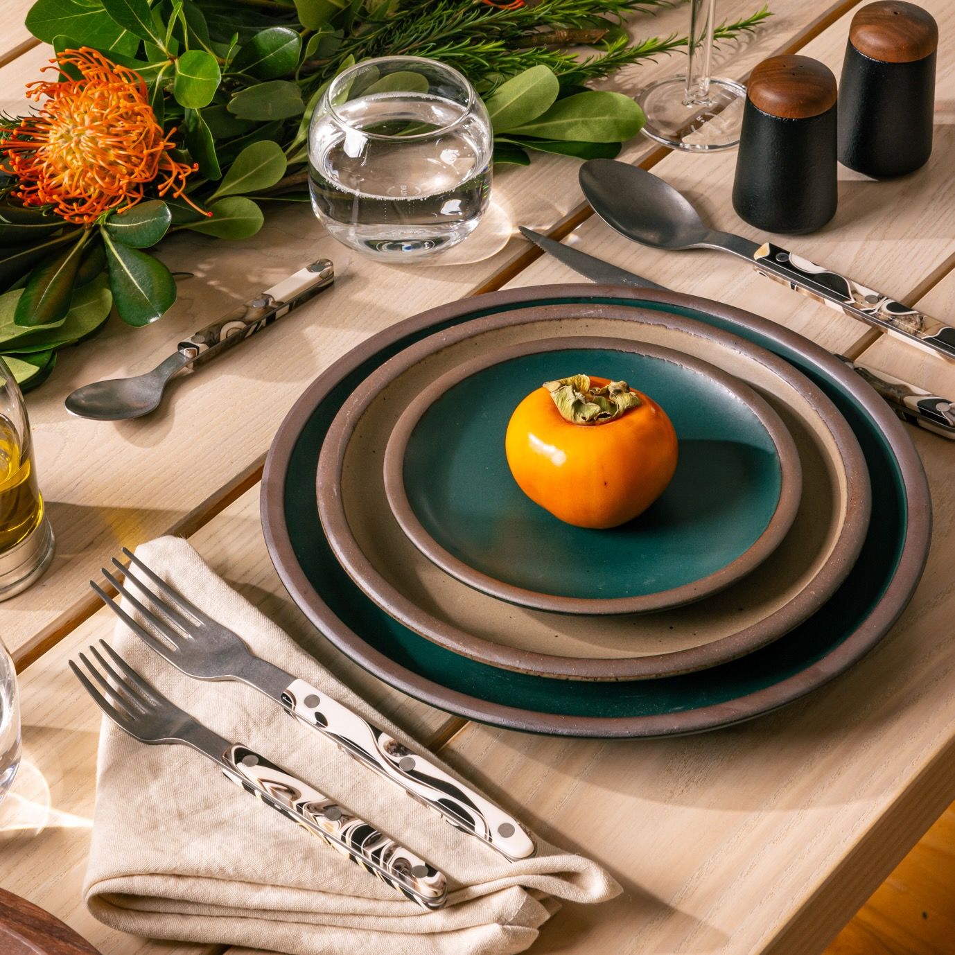 A place setting featuring dark teal and natural colored ceramic plates with black, white and cream marble handled cutlery (salad fork, dinner fork, knife, soup spoon and teaspoon), Also pictured is a folded neutral dinner napkin, an a rounded stemless glass filled with water.