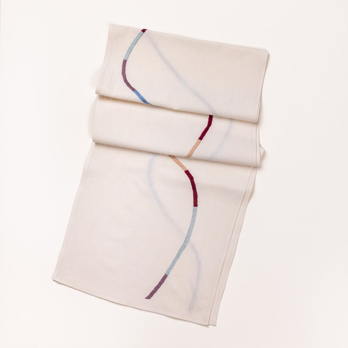 A folded table runner in a natural cotton with a thin embroidered line curving down the runner in color blocks of purple, peach, and periwinkle colors