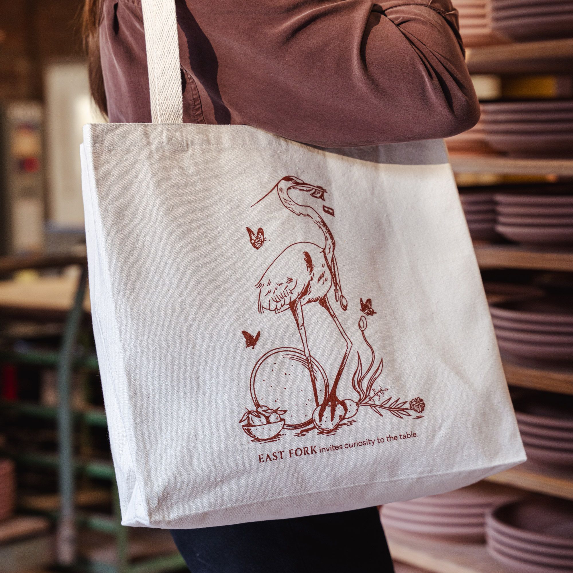 A model wearing a tote with an illustration of a heron with ceramic plates and text that reads 'EAST FORK brings curiosity to the table.'