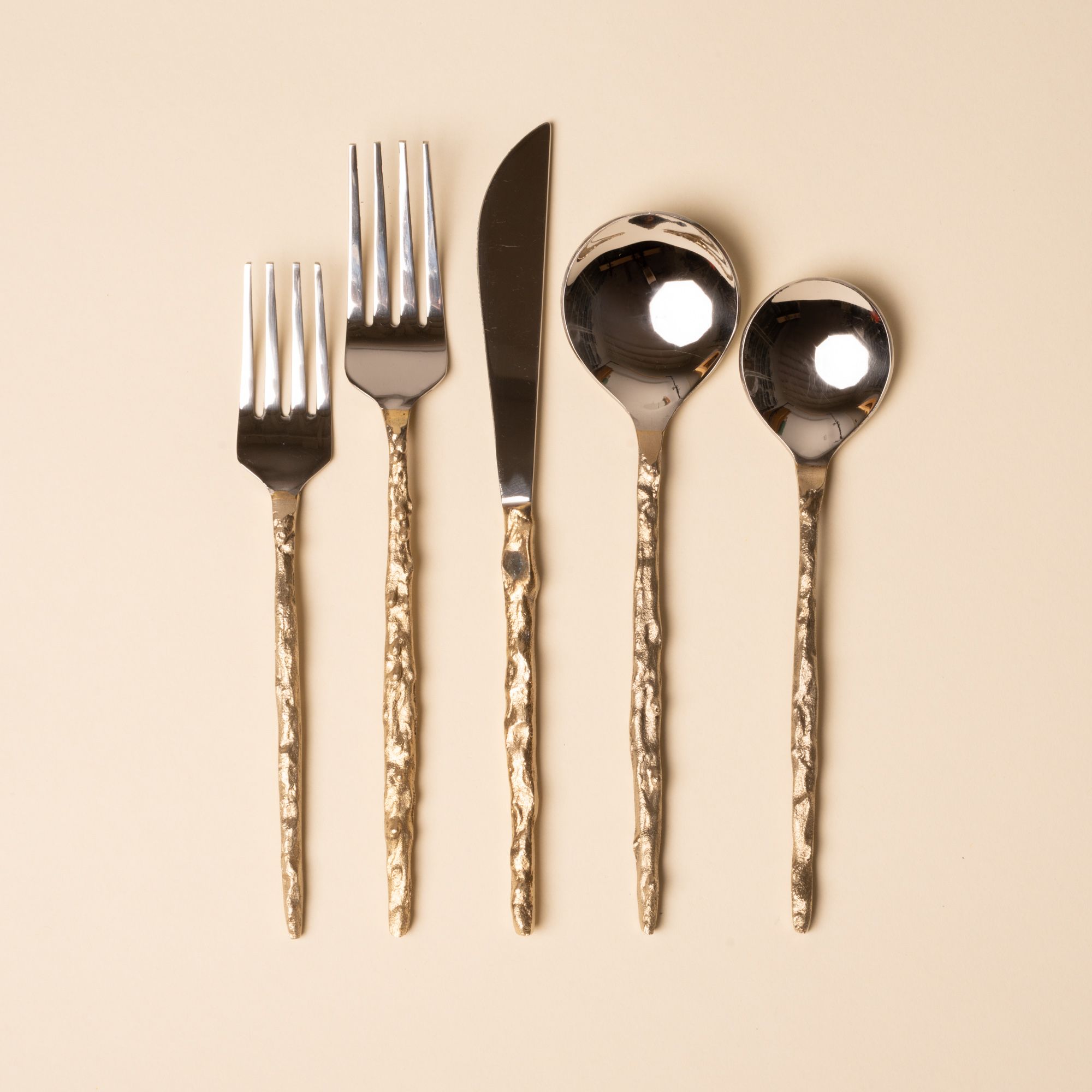 Shiny silver flatware set with hammered gold handles arranged on a beige background.