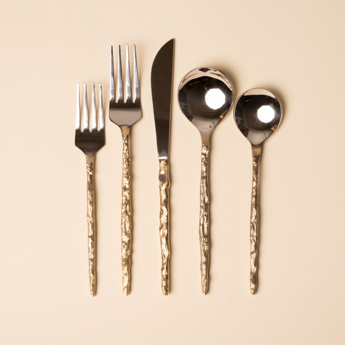 Shiny silver flatware set with hammered gold handles arranged on a beige background.