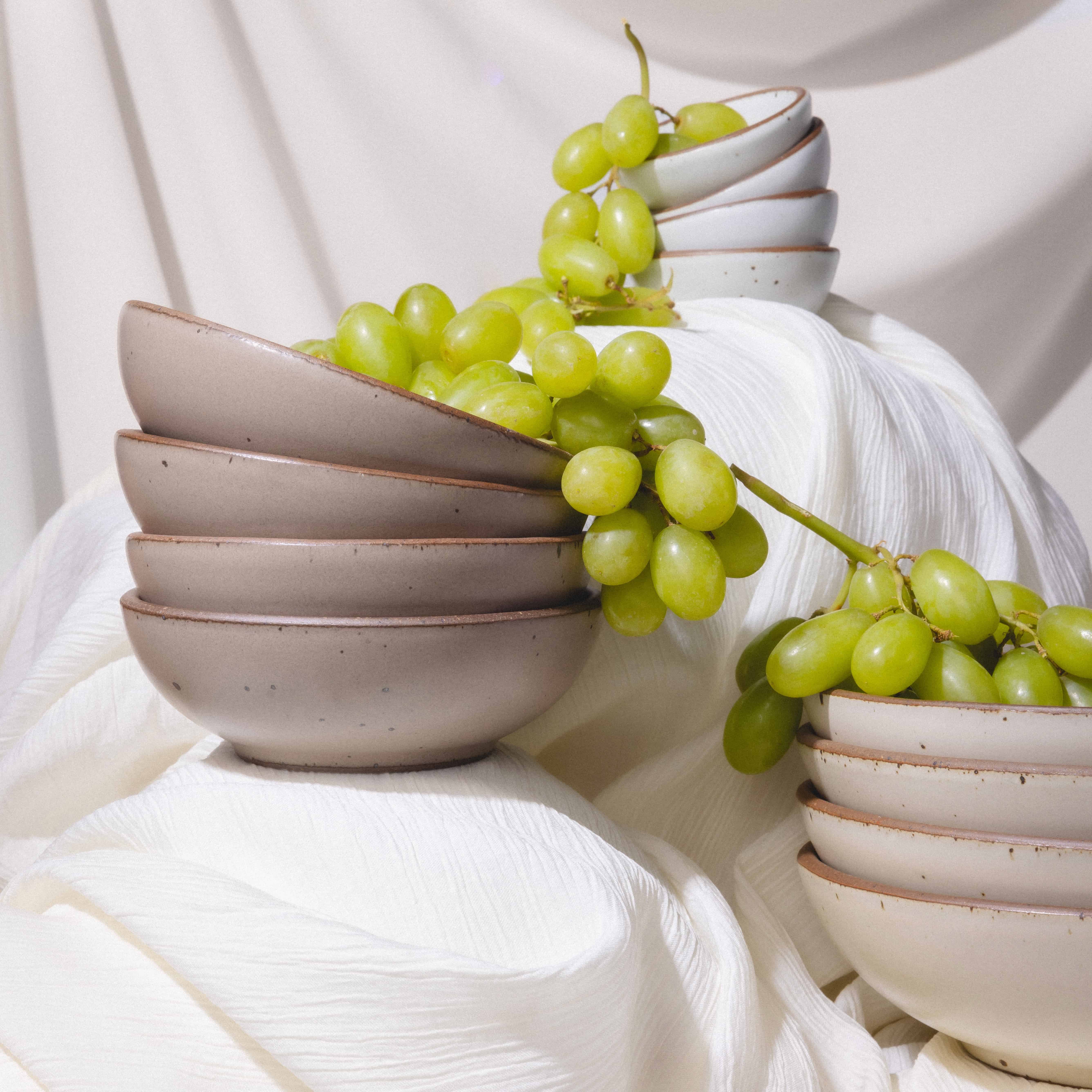 Stacked bowls in various sizes in neutral colors with green grapes styled on top with white drapery.