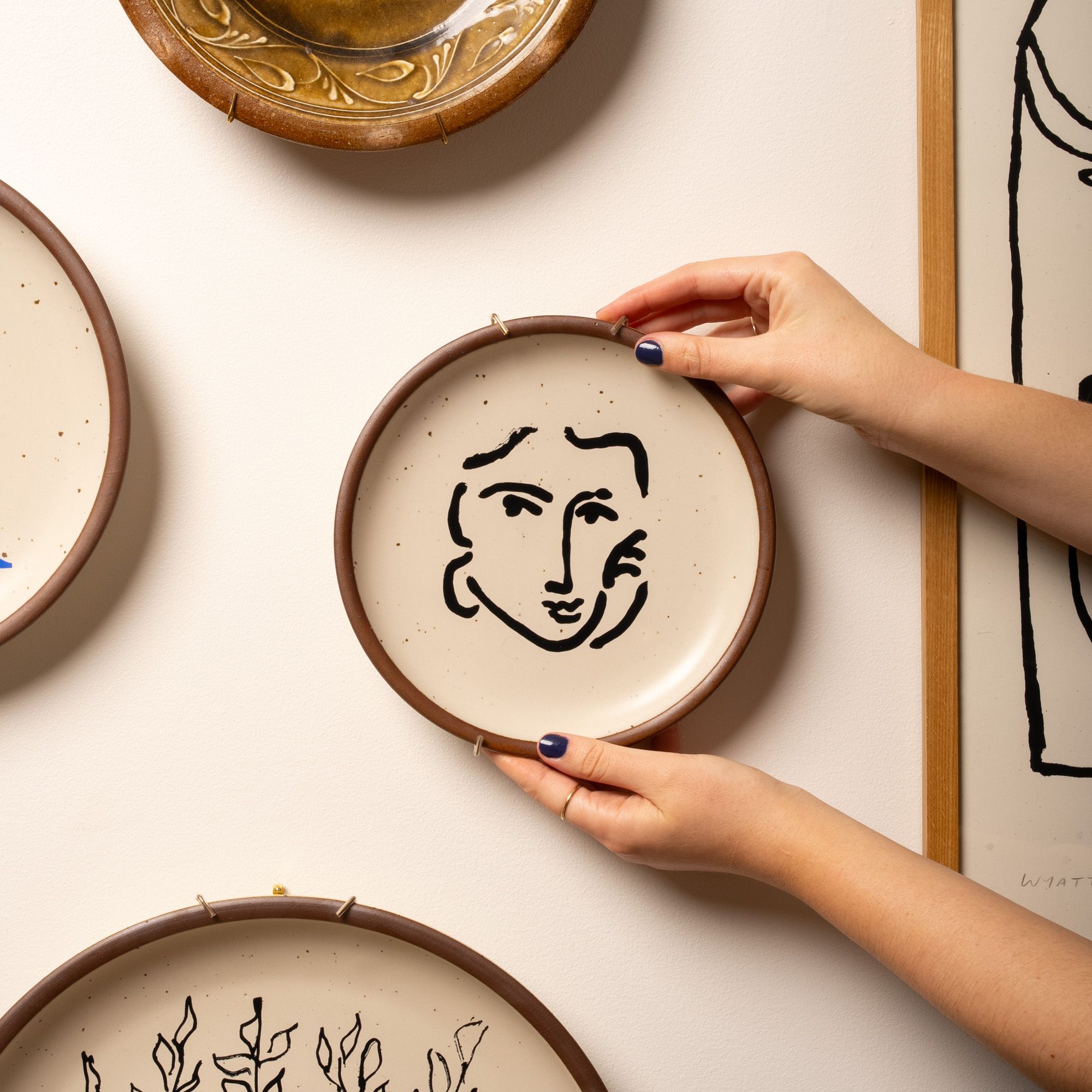 A hand holds up a side plate with an artful face decal on it, against a wall to hang it up.
