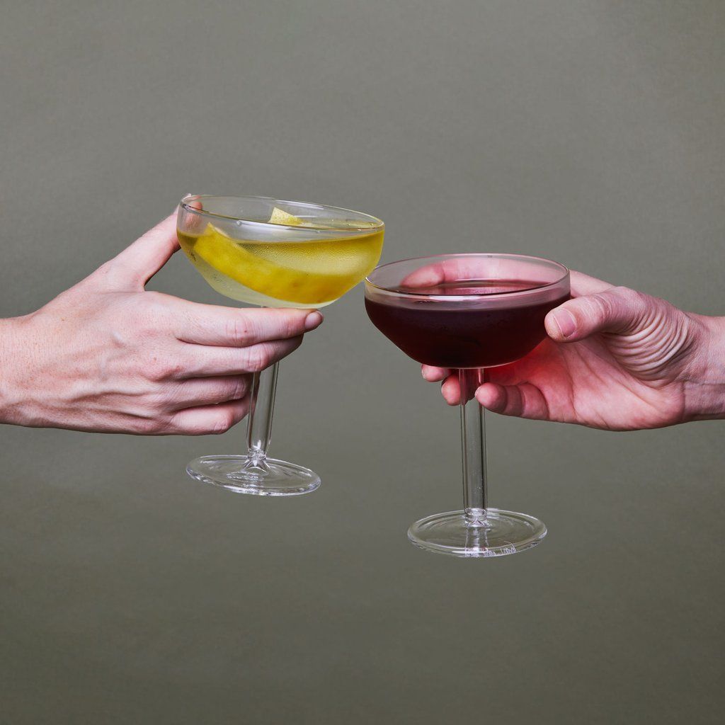 Two hands each hold a Martini coupe, one filled with clear liquid and a twist of lemon and the other filled with red wine