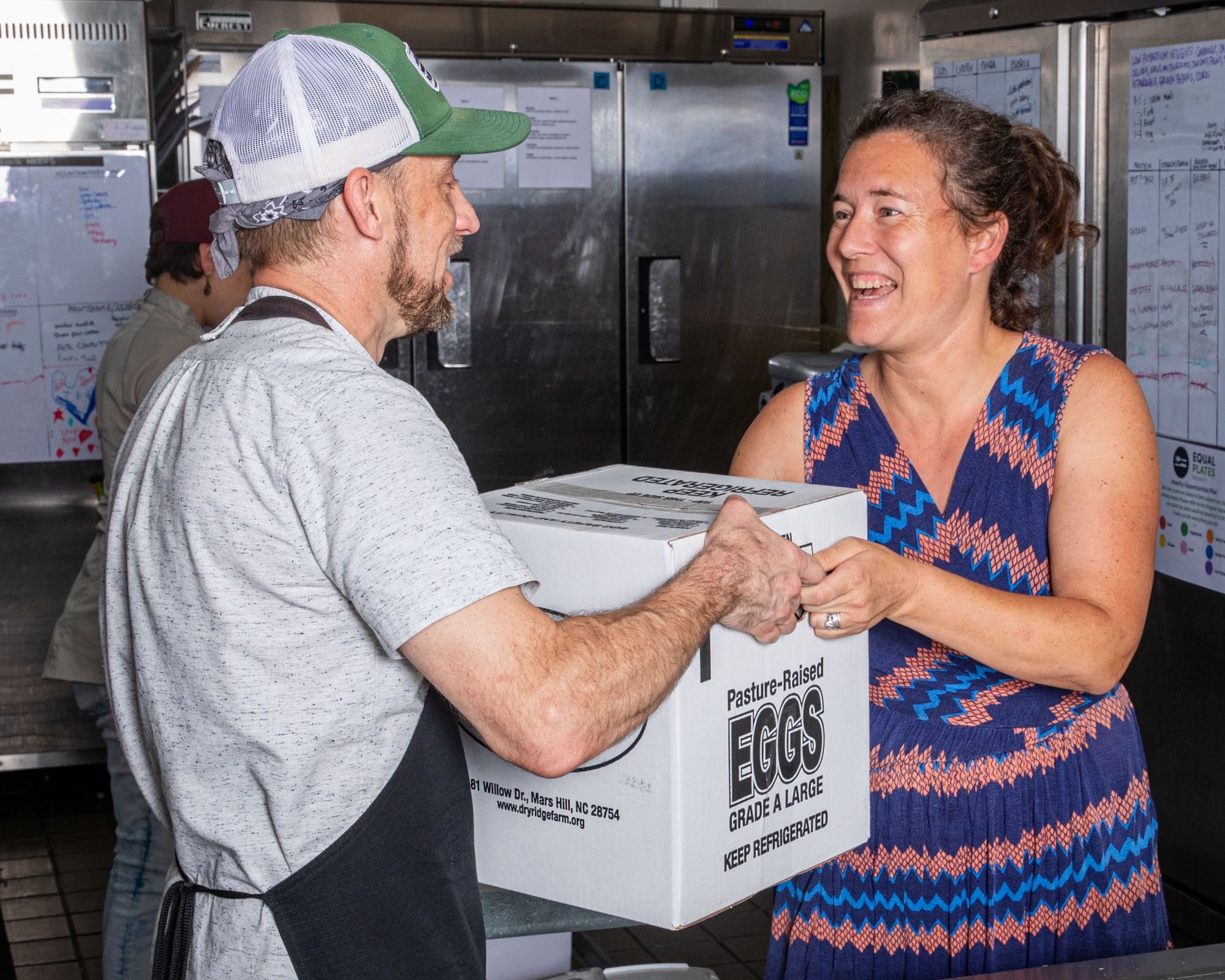 In a kitchen, a person hands a box of eggs to another person and is smiling.