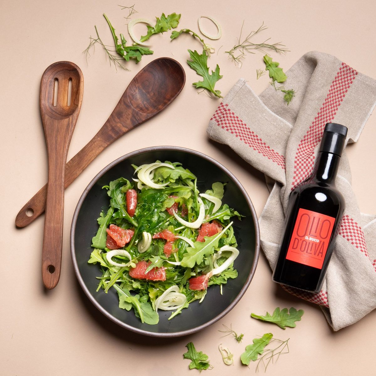 A large black ceramic bowl filled with arugula, fennel, and grapefruit sits beside olive oil, a red-striped towel, and wooden salad servers.