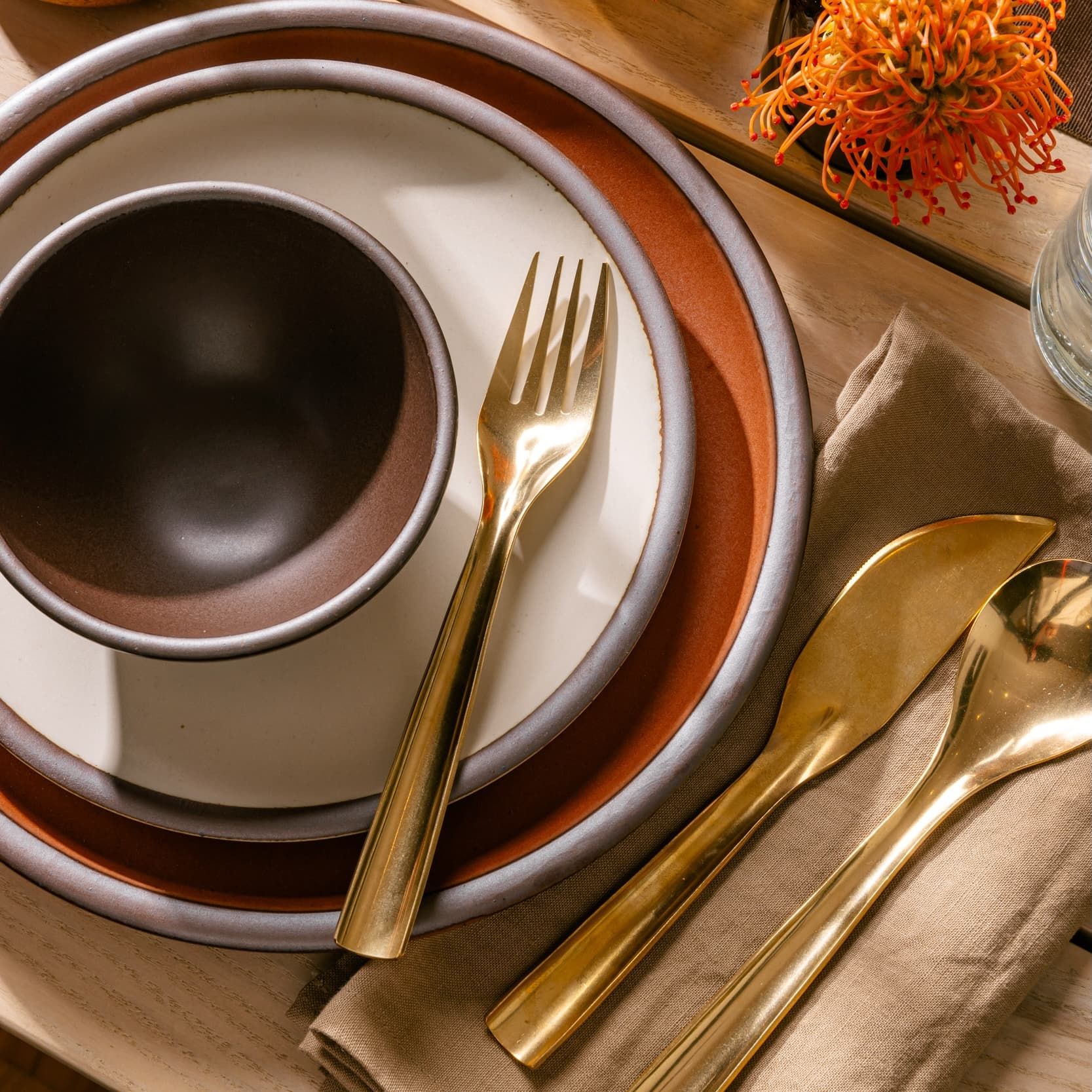 On a table is a stack of plates and bowl in cool terracotta, cream, and dark brown colors. To the right is a shiny brass fork, knife, and spoon on a natural linen napkin.