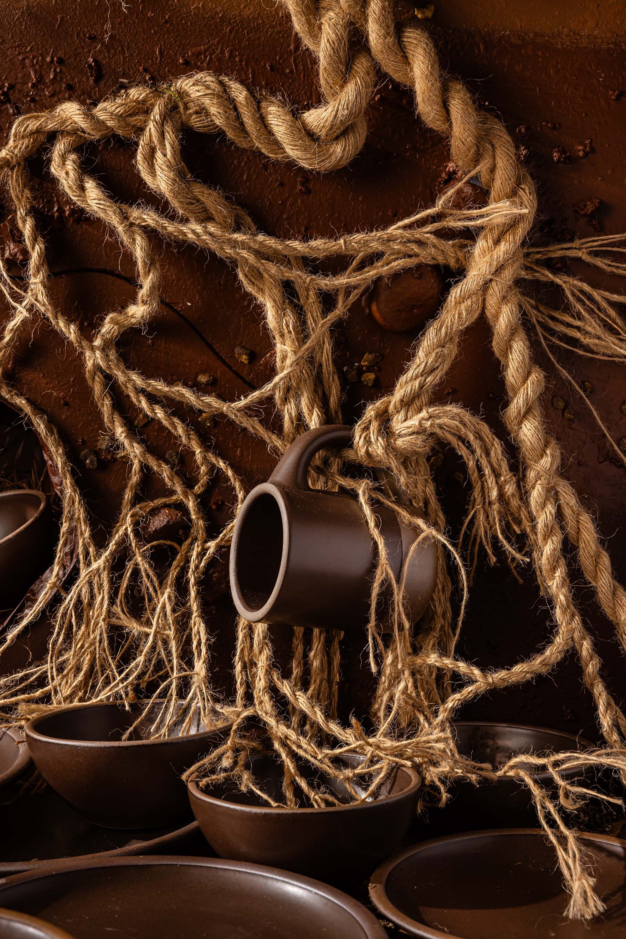 Various plates, bowls, and a mug in a dark brown all surrounded by ropes and dirt.