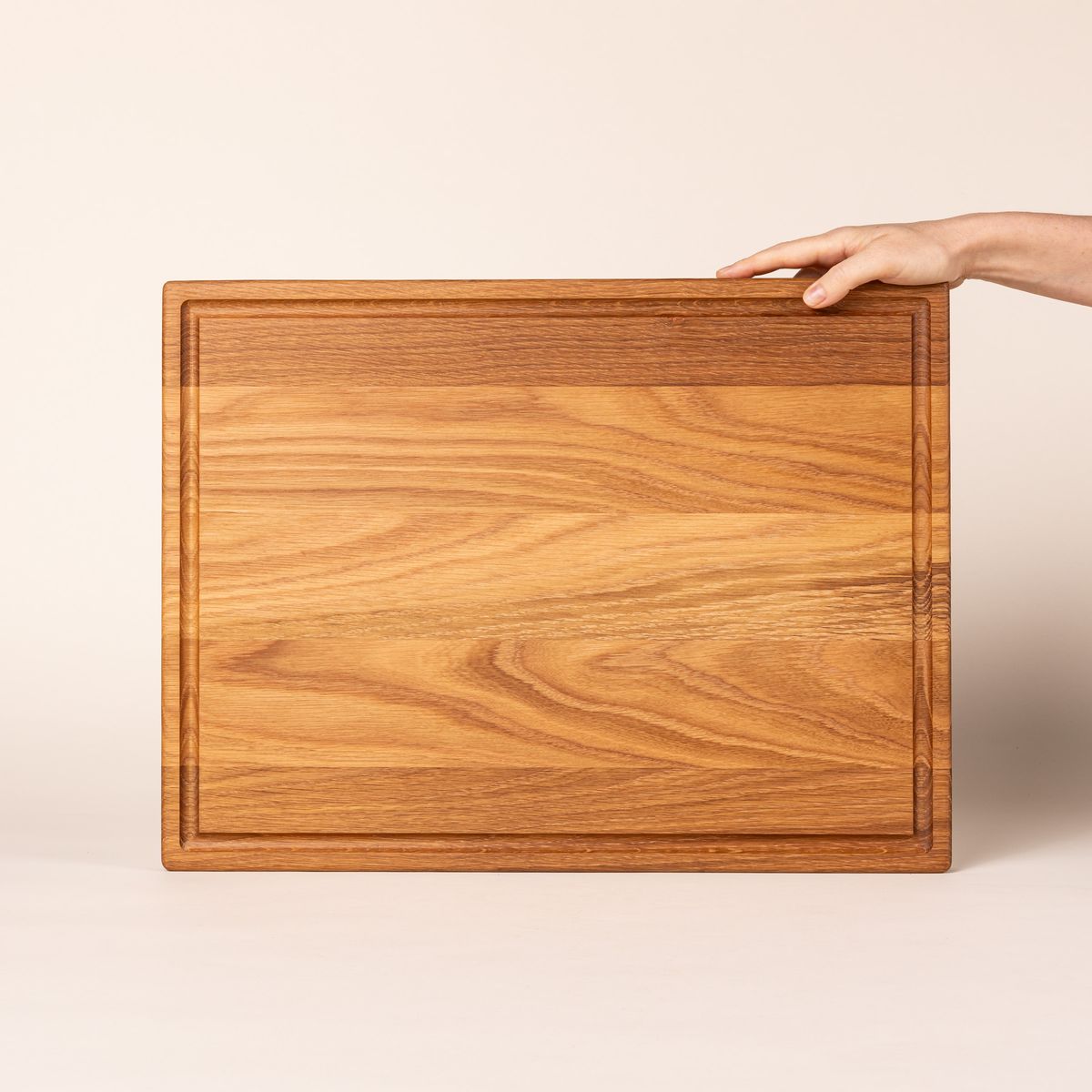 A hand props up a large rectangular white oak cutting board on its side with a juice groove