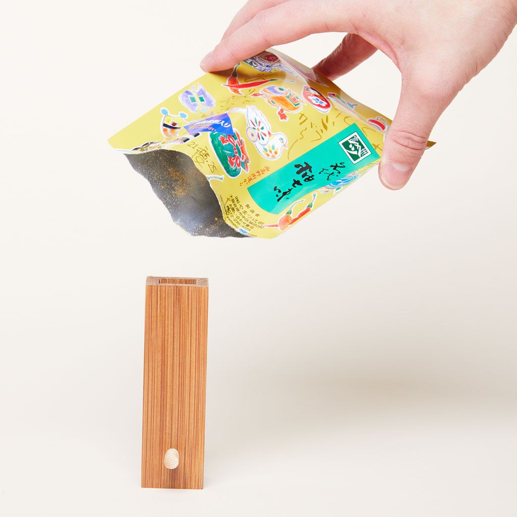 A hand holds a colorful bag of spices above a wooden rectangular container that is open at the top