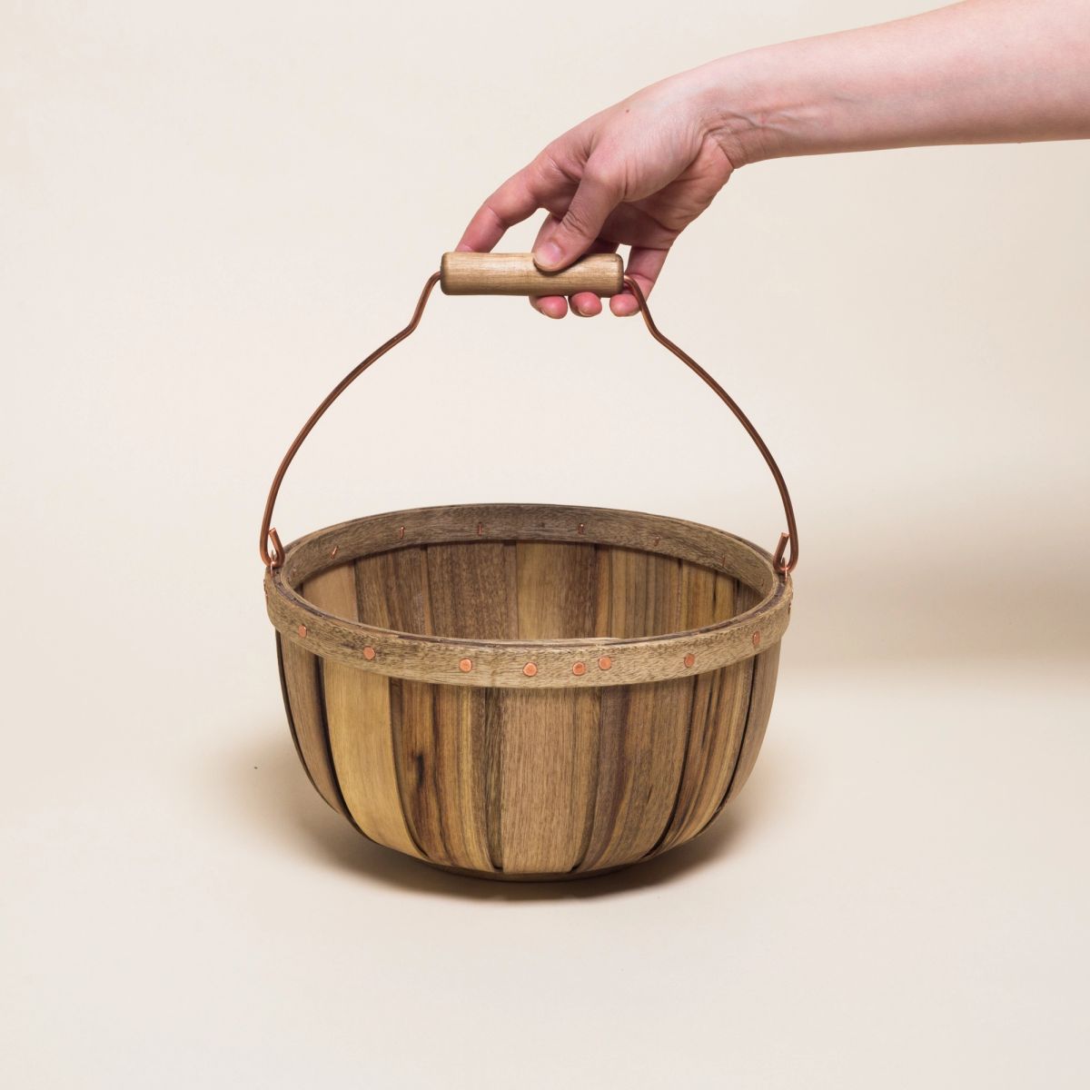 A hand holds the metal handle of a round wooden basket