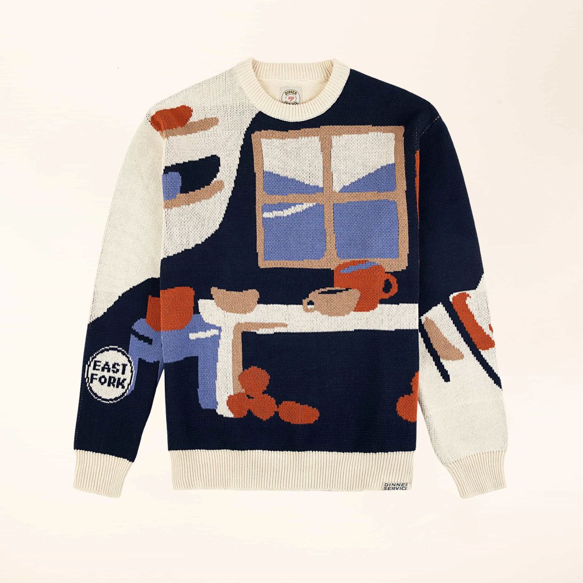 A flatlay of a sweater with an illustration of a pottery workshop with a large window, shelves, and table with mugs and bowls. The sweater features cream, navy, terracotta, tan and blue colors.