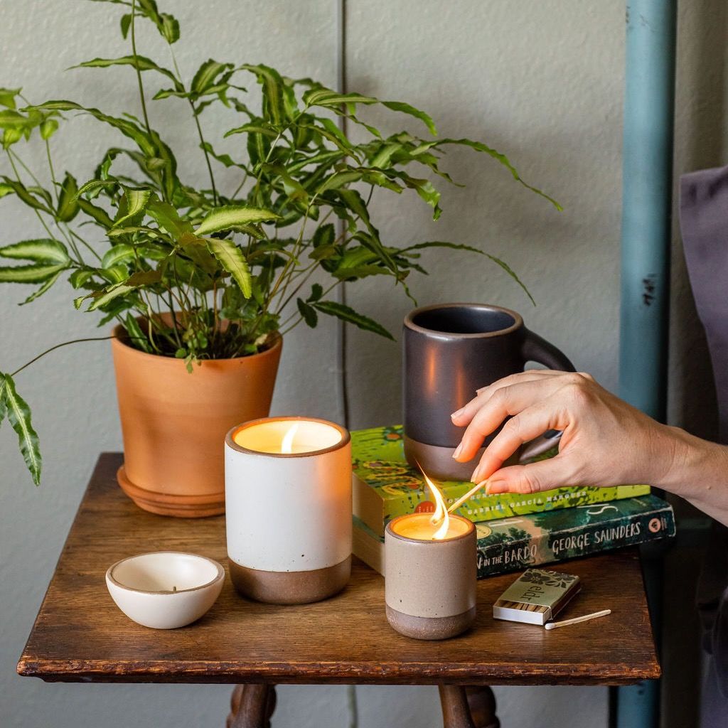 Ceramic candles are sitting a bedside table next to a box of matches, houseplant, and mug. A hand is lighting the smaller candle.