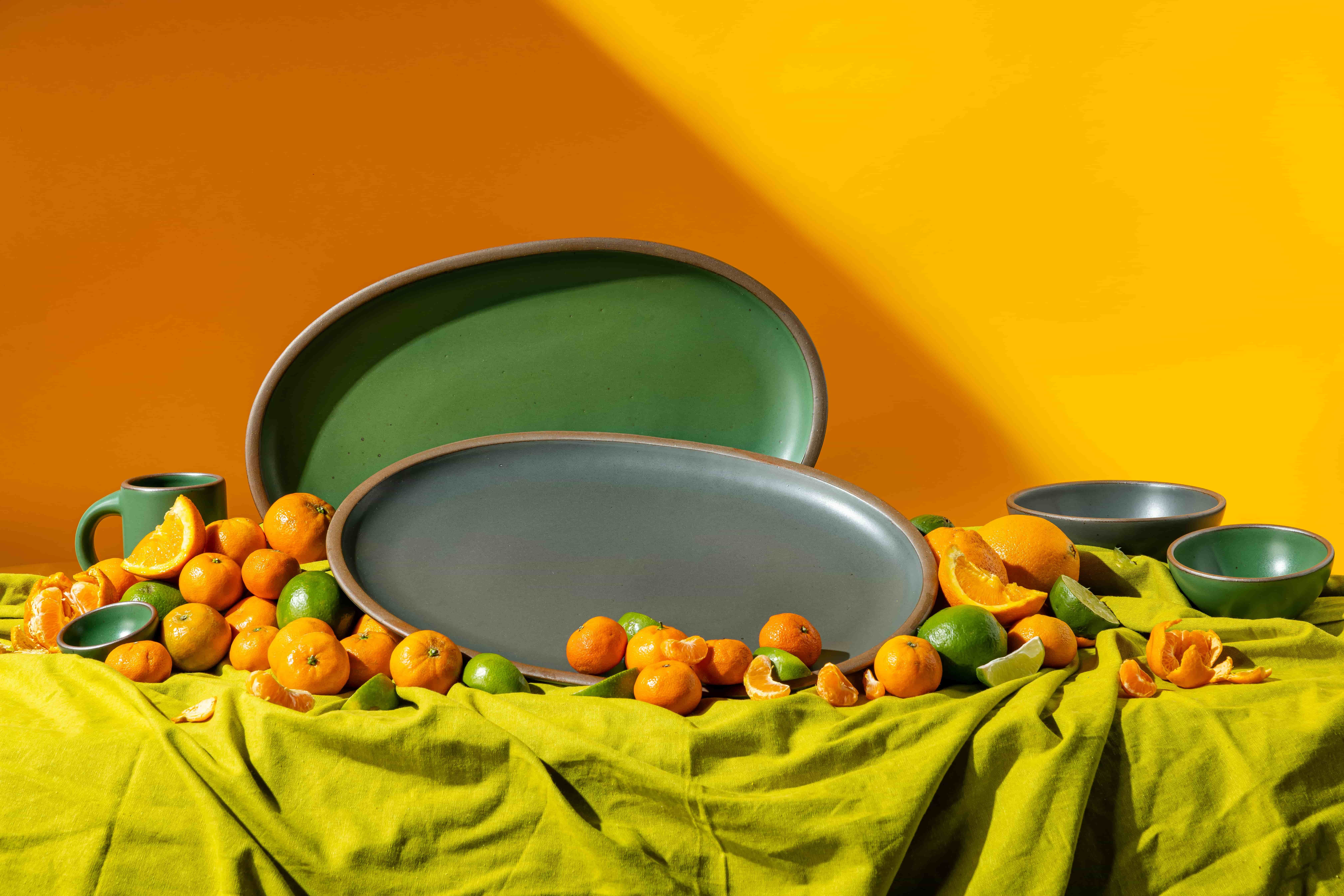 Two ceramic oval platters in green and grey colors on a table surrounded by bowls in the same color and lots of oranges and limes