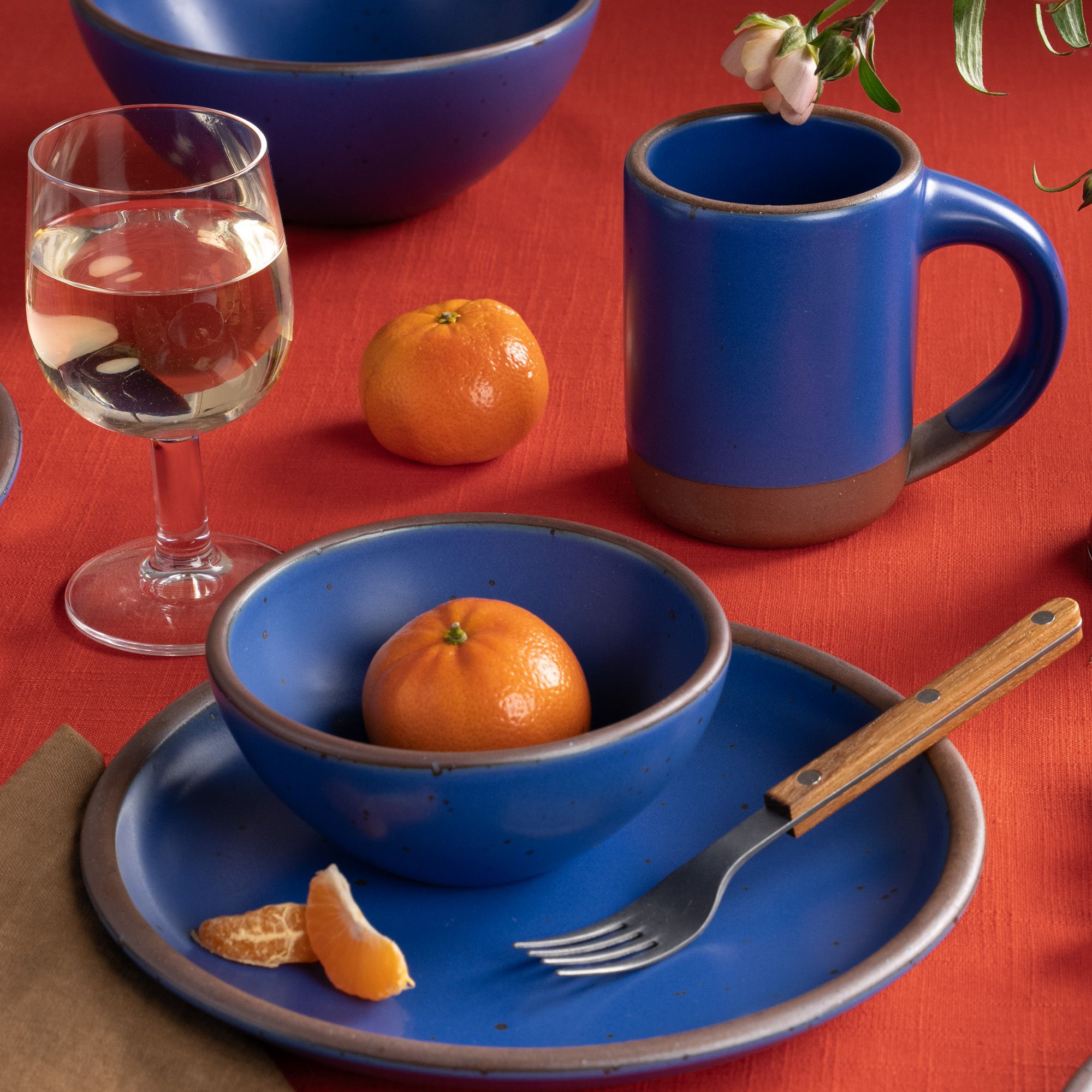 Blue ceramic dinnerware with clementines, a fork, and a mug on a red tablecloth beside a glass of white wine.