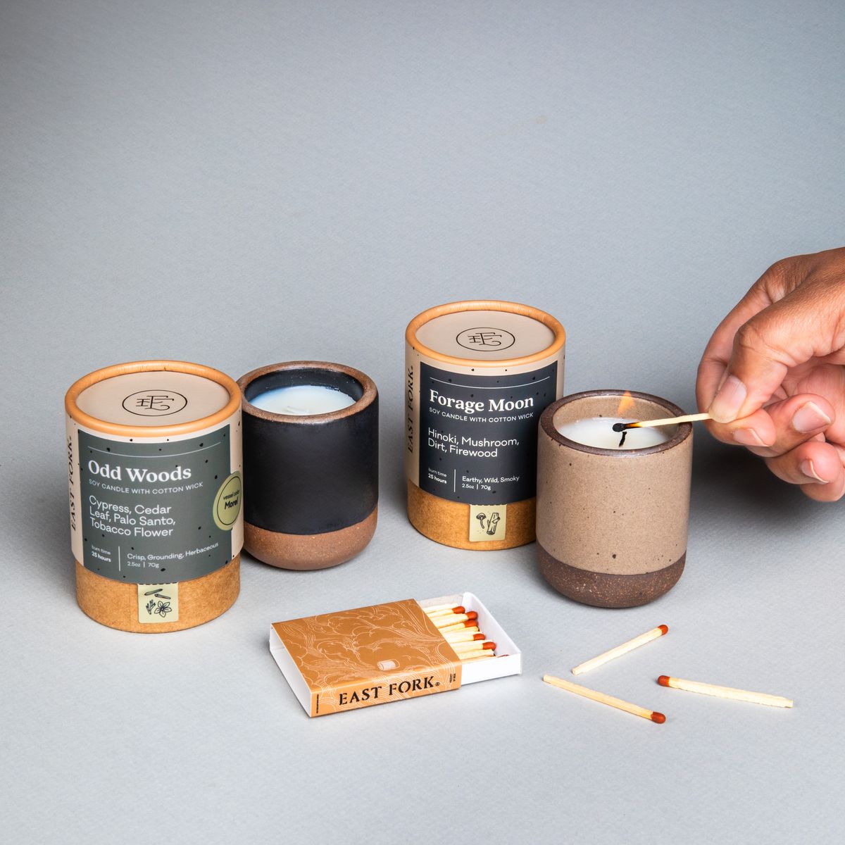 Two small ceramic vessels in dark charcoal and warm pale brown colors with candles inside each are gathered next to their cardboard packaging tubes with matches. A hand is lighting the warm brown candle.