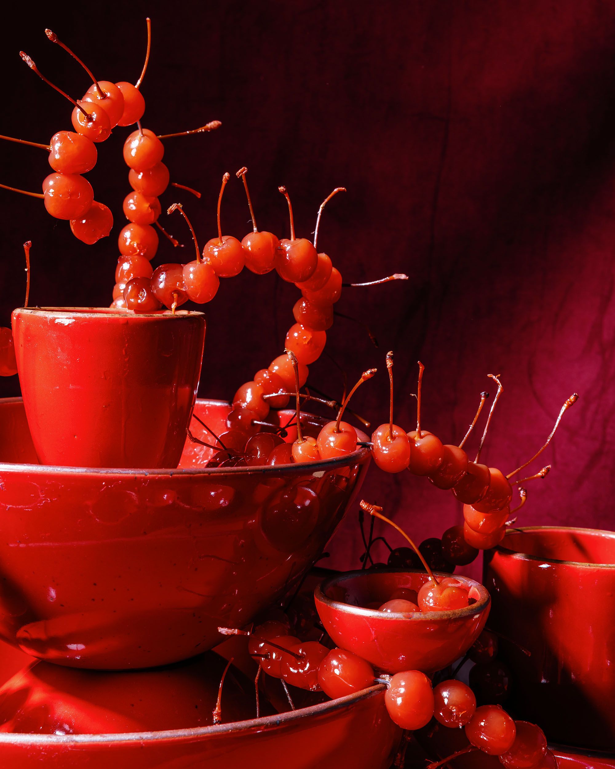 Glossy red bowls and a cup entwined with strands of bright red cherries against a deep red background.