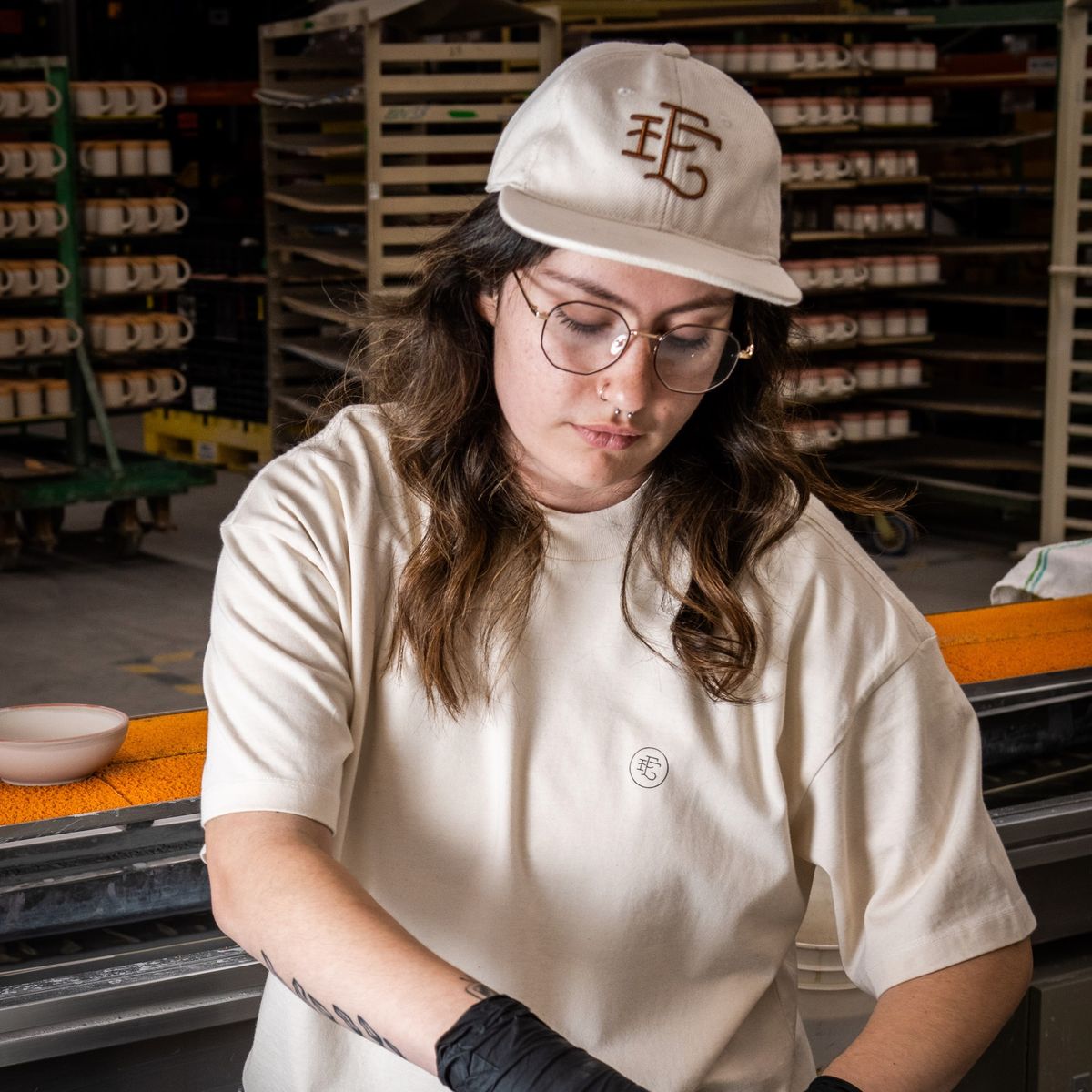 In a factory warehouse setting, a person wears the white denim baseball hat while handling a ceramic bowl on the production line.