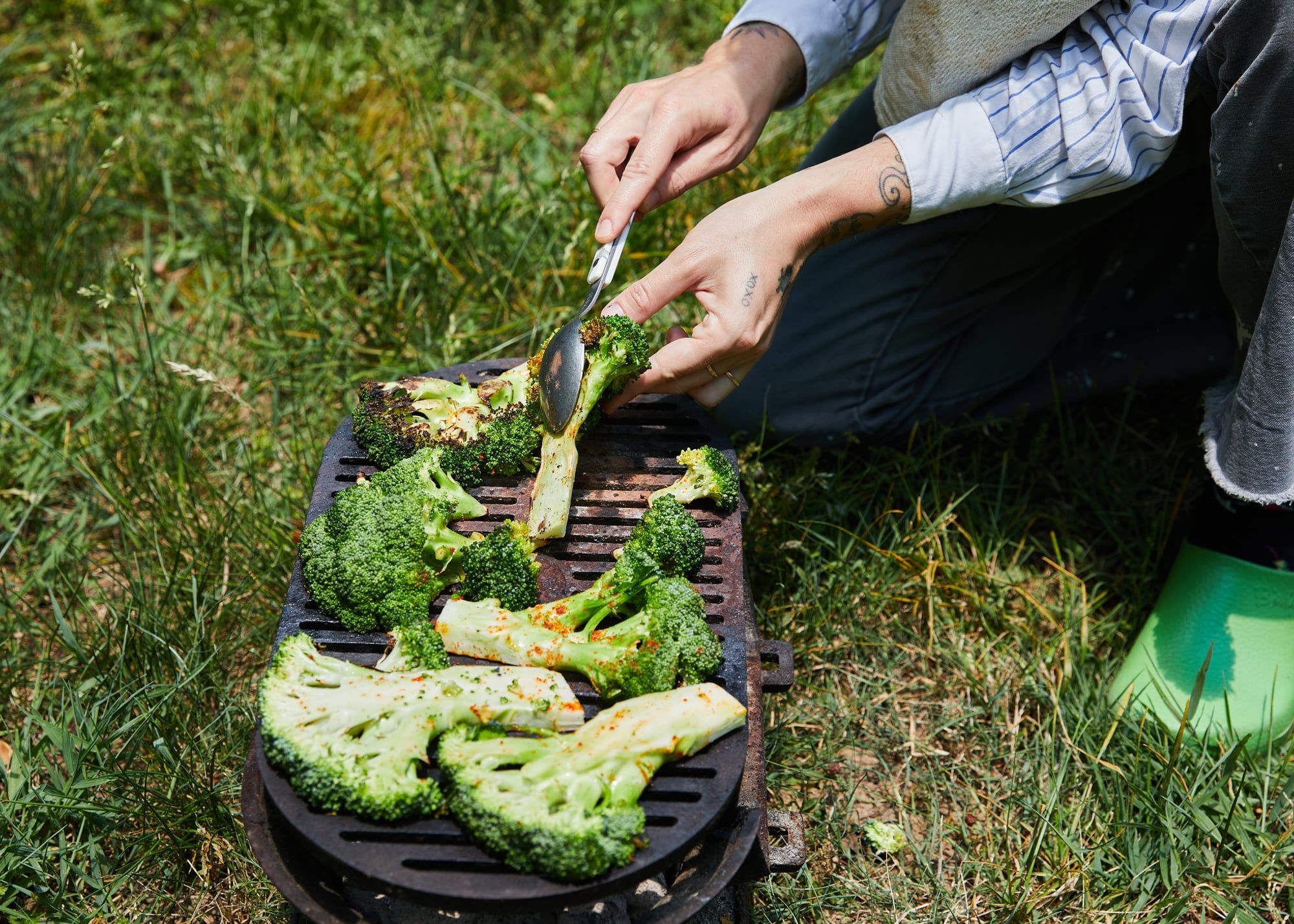 A person is charring broccoli outside on the grass with a flat grill