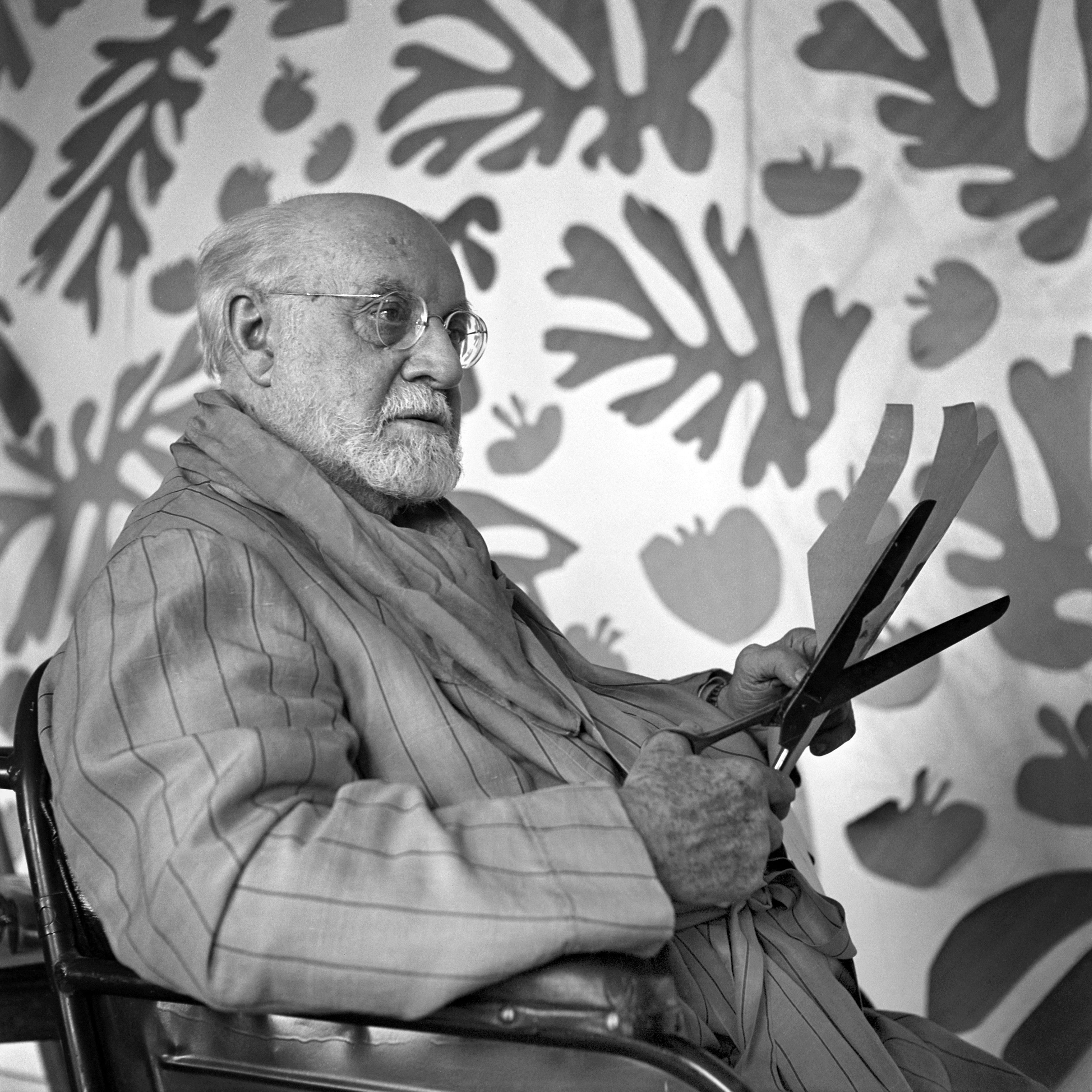 Black and white photograph of Henri Matisse sitting in his studio with a pair of scissors and a large wall of artwork behind him.