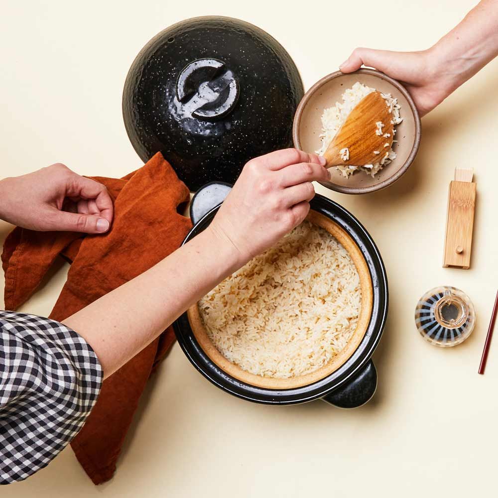 Birds-eye-view of a table where a hand is serving rice from a Japanese clay pot donabe with a wooden rice spoon into a stoneware bowl. A soy sauce cruet and bamboo spice container are also on the table.