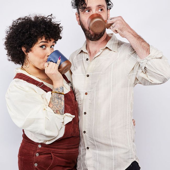 Two people are holding each other and each carrying an East Fork mug up to their face. The left person is holding a Blue Ridge, or muted navy color mug and looking at the camera, and the person on the right is drinking out of a Amaro glaze mug, a cool terracotta color.