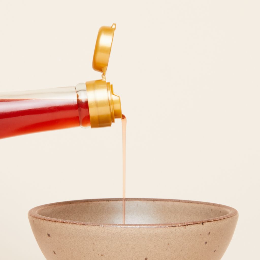 A thin stream of purple sweet potato vinegar pours from a glass bottle with a gold flip-top cap into a brown bowl