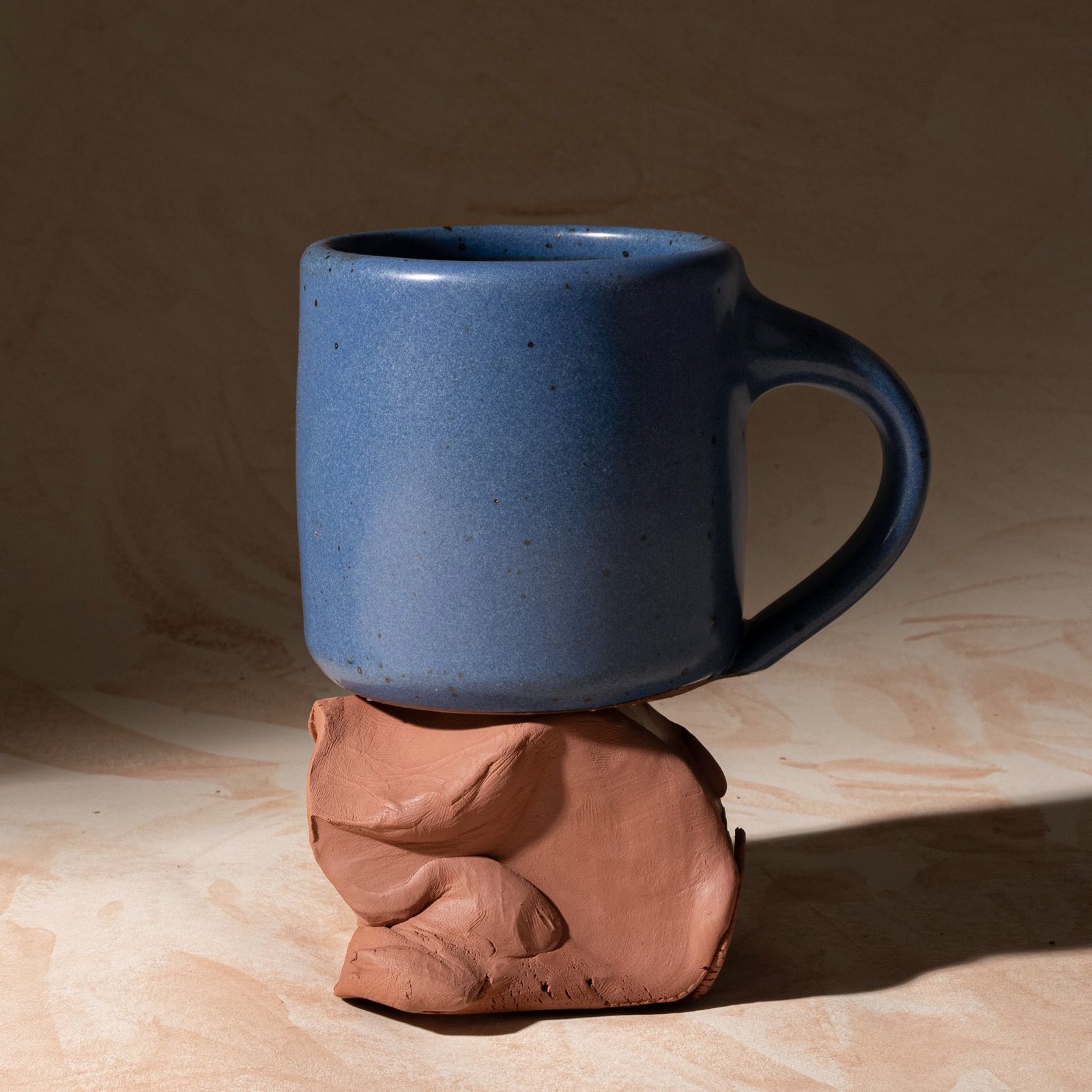 A medium sized ceramic mug with handle in a cool, medium blue color featuring iron speckles, sitting on a block of raw clay.