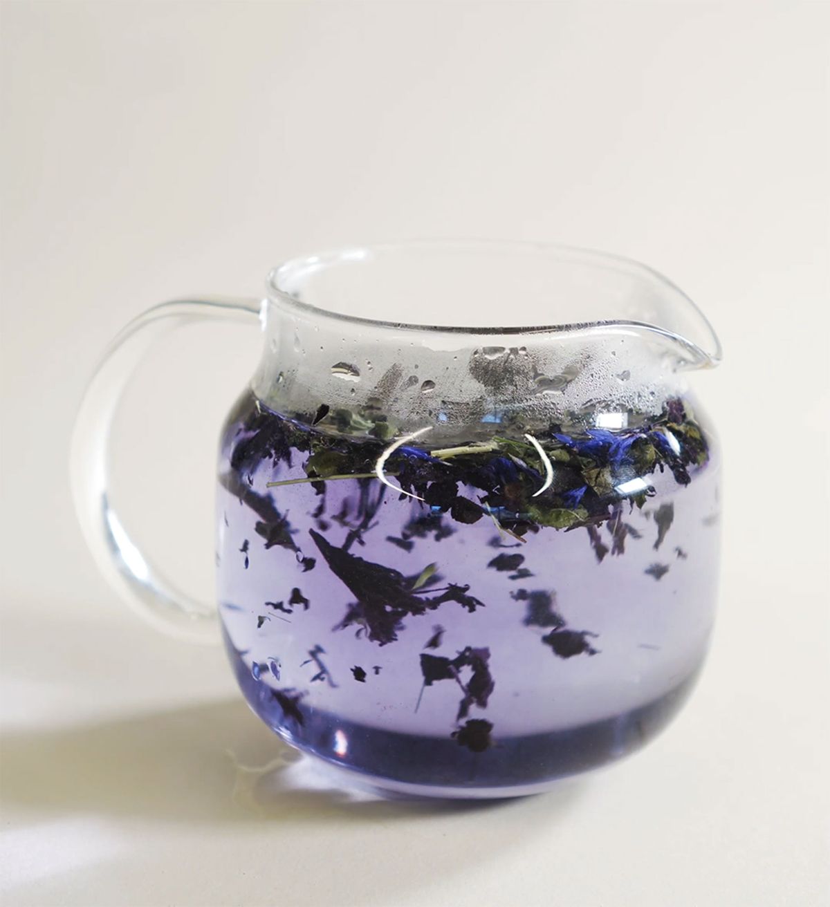 Glass teacup with spout holding light purple liquid and blue and green tea leaves inside.