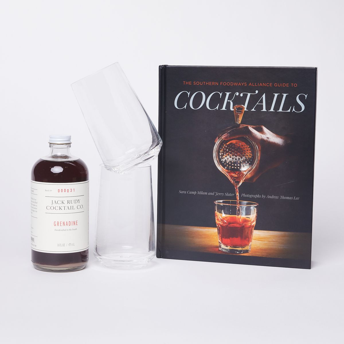 A full bottle of grenadine next to a set of stacked clear glass tumblers and a book with a cover the shows a picture of a drink being poured with the word "Cocktails"