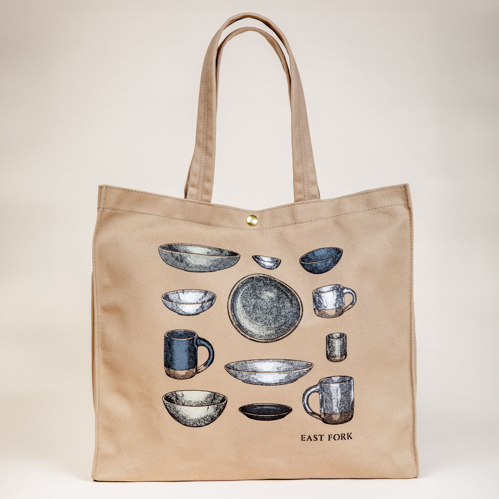 A sturdy canvas tan tote bag with handles and a metal closure button at the top. On the bag is a printed illustration in neutral colors of bowls, plates, and mugs.