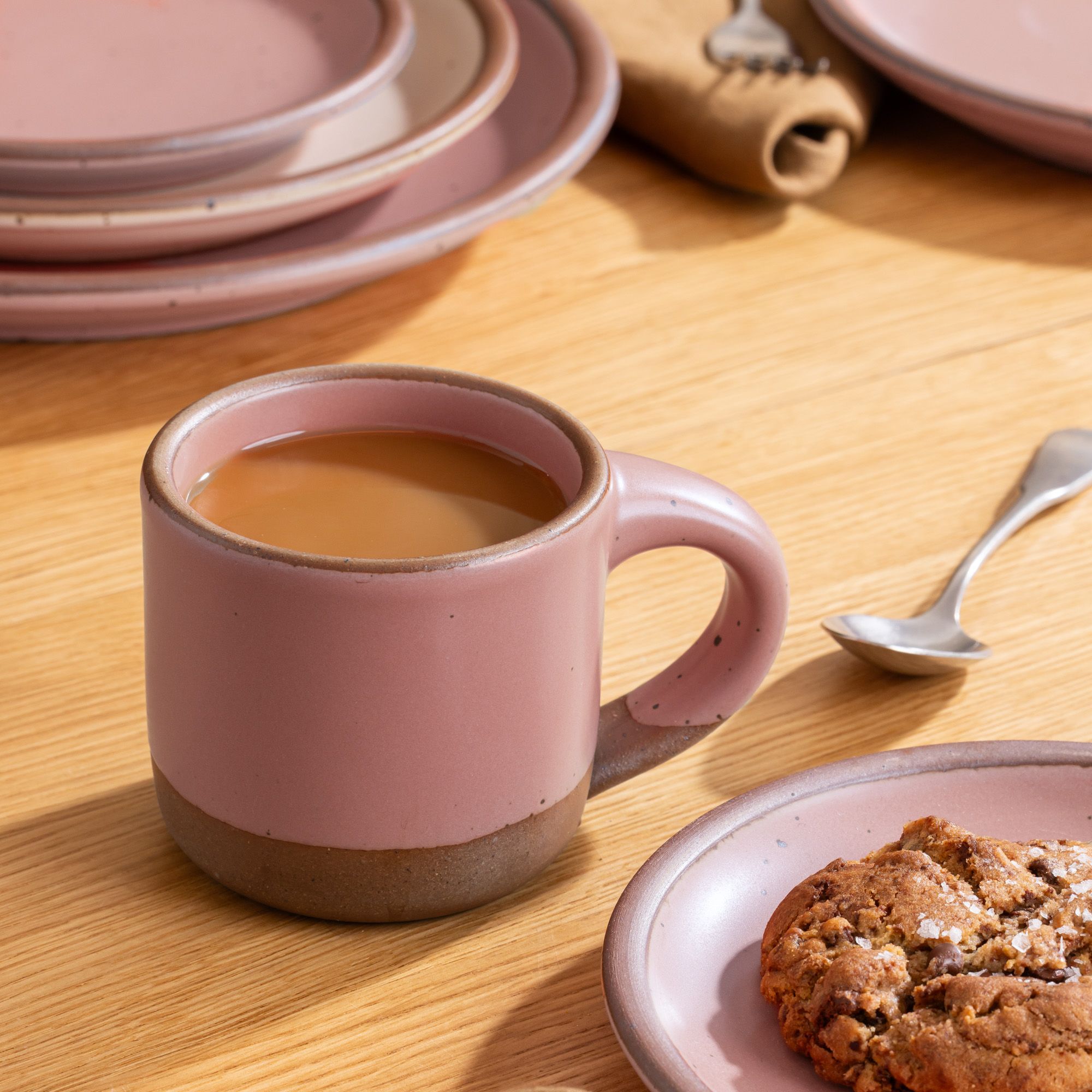Dusty mauve pink ceramic mug filled with coffee beside a cookie on a matching plate, set on a wooden table with dishware in the background.