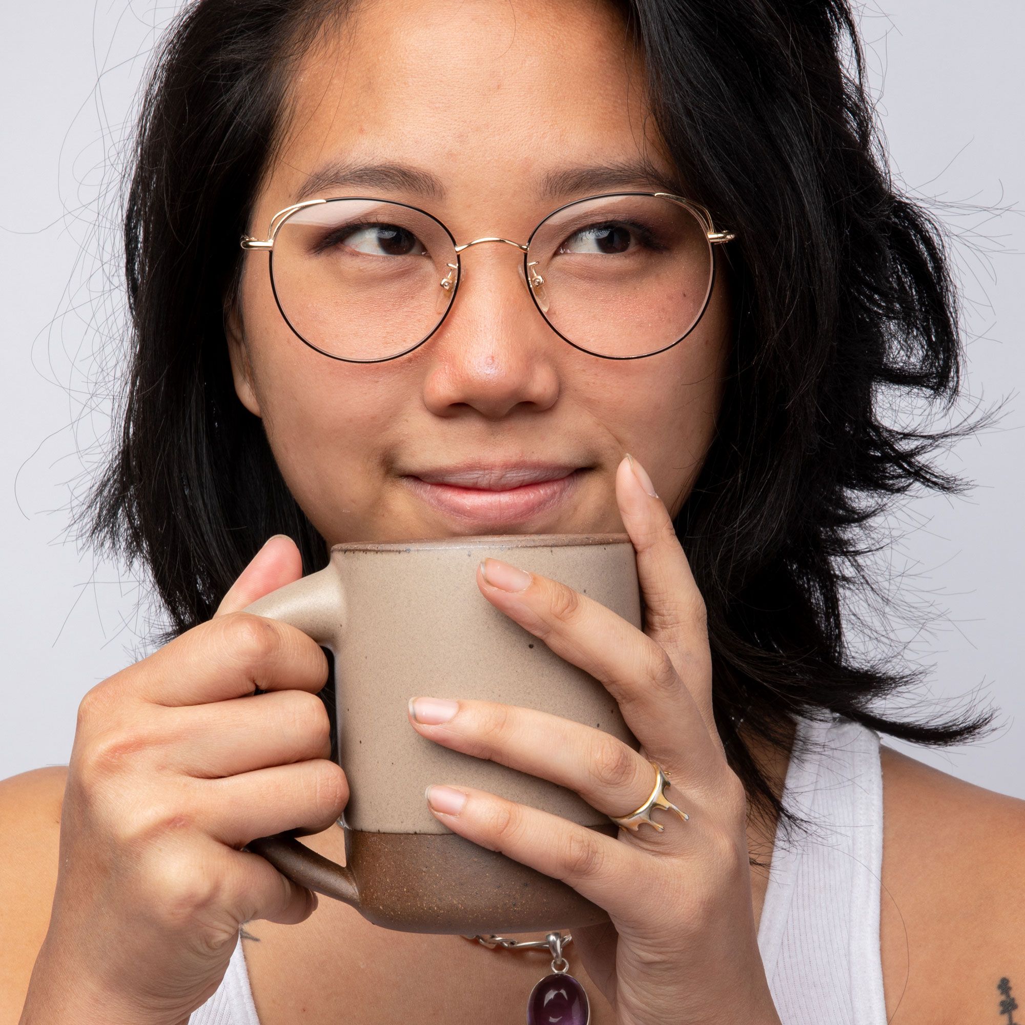A person holding a medium sized ceramic mug close to their face. The mug is in a warm pale brown color featuring iron speckles and unglazed rim and bottom base.