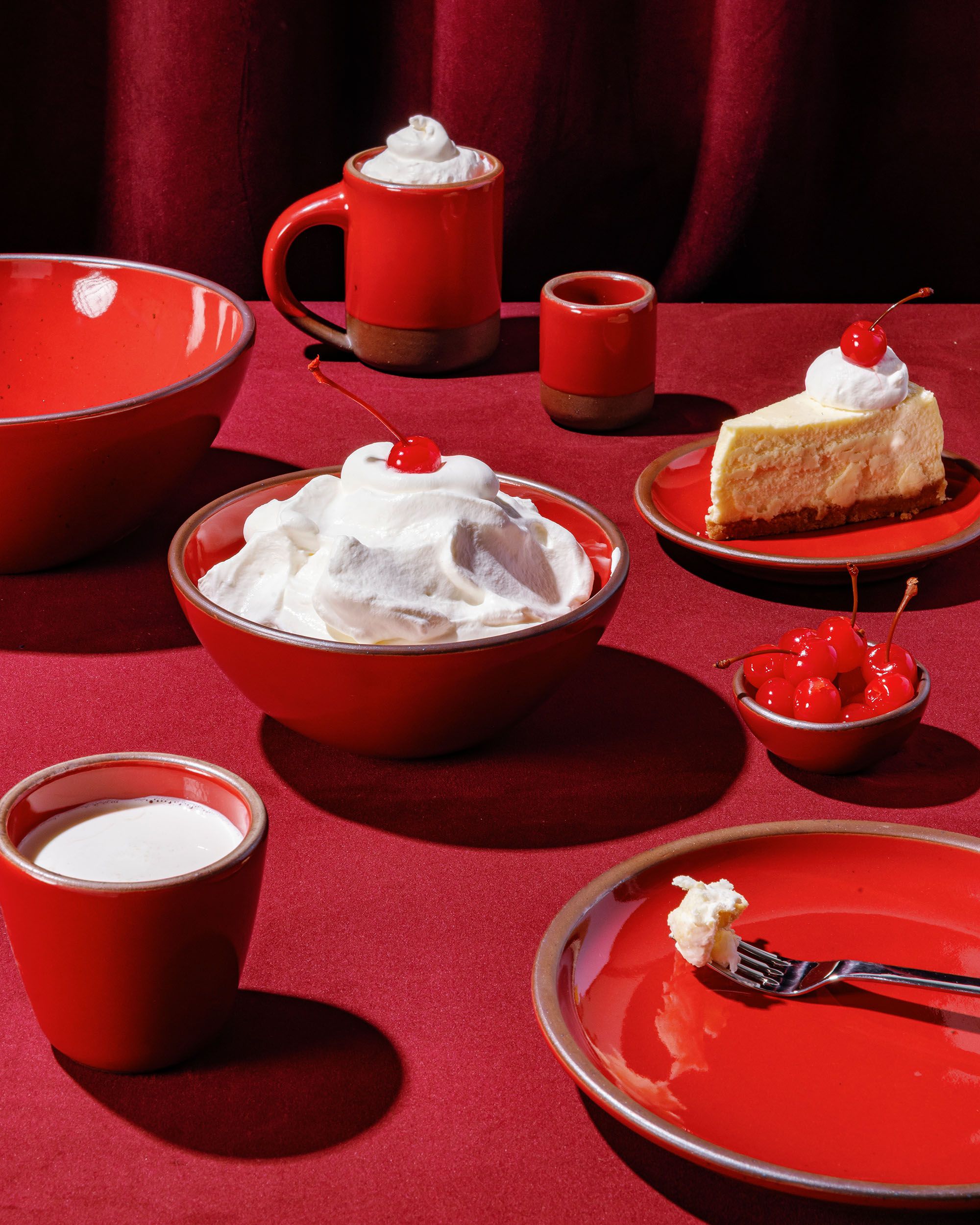 Glossy red ceramic bowls, cups, and plates with whipped cream, cheesecake, and cherries, against velvet red fabric background.