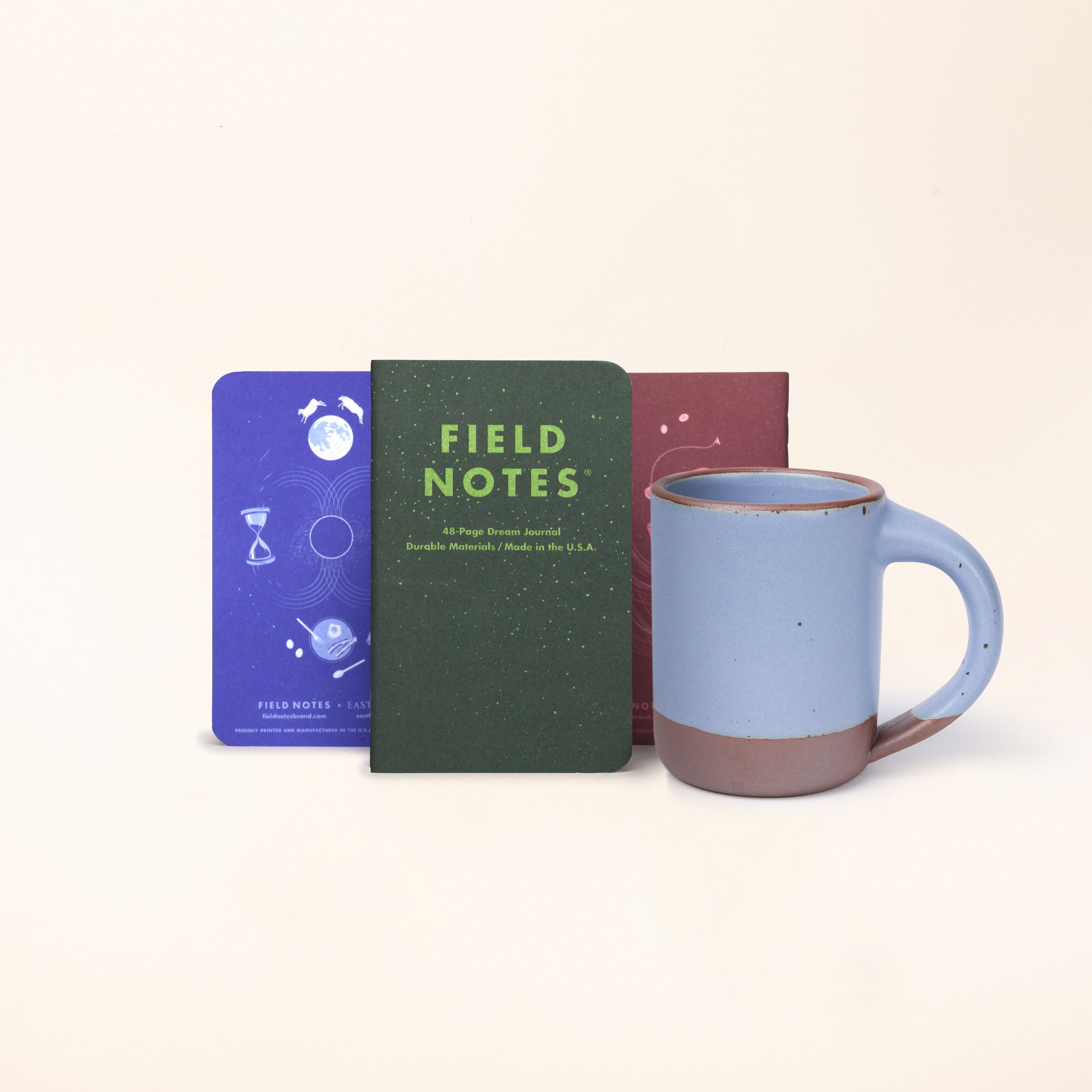 A 3 pack of Field Notes Journal standing up in a row in forest green, lapis blue, and plum colors with The Mug in a periwinkle color.