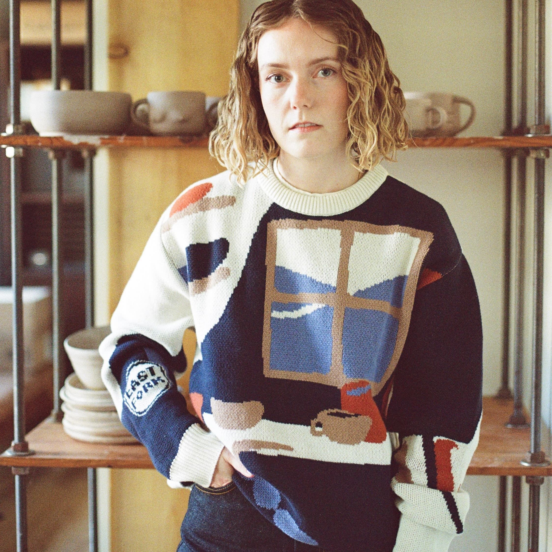 Erica has a hand in her pocket and wears a sweater with an illustration of a pottery workshop with a large window, shelves, and table with mugs and bowls.