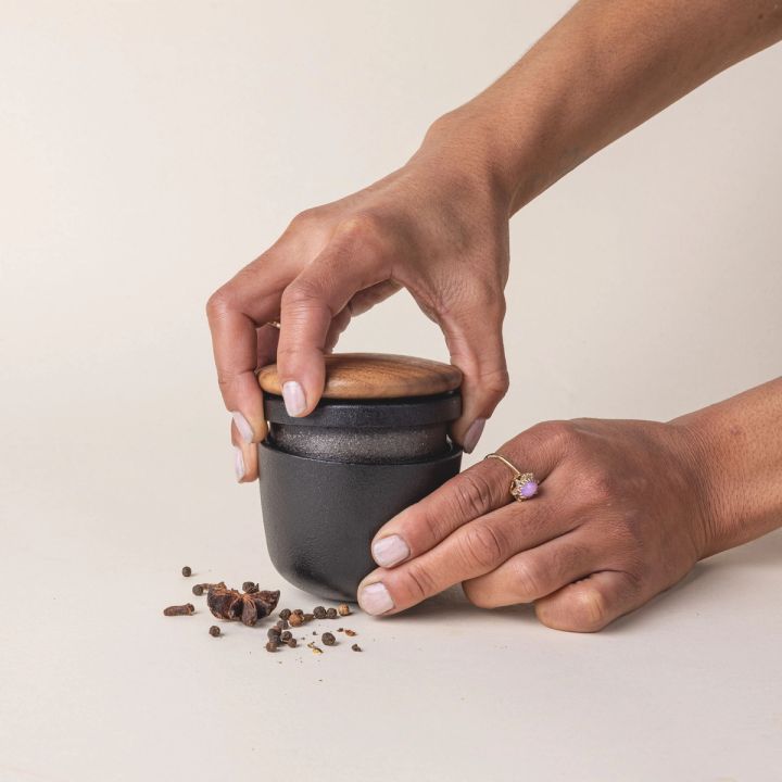 Hands hold a short round cast iron jar with a topper grinder, grinding a seasoning inside the jar.