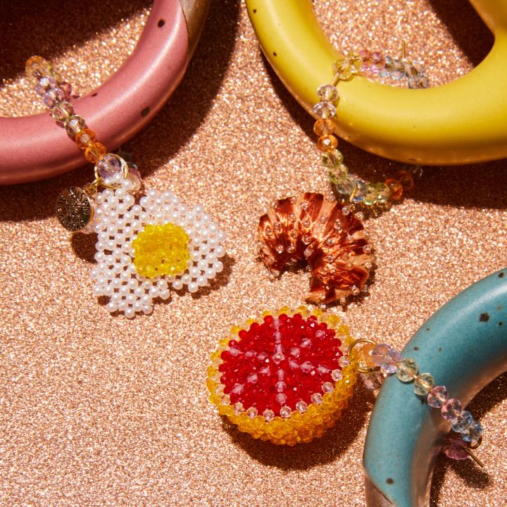 1 beaded egg mug charm attached to a handle of a pink mug, 1 beaded croissant mug charm attached to a handle of a yellow mug, 1 beaded grapefruit mug charm attached to a handle of a turquoise mug