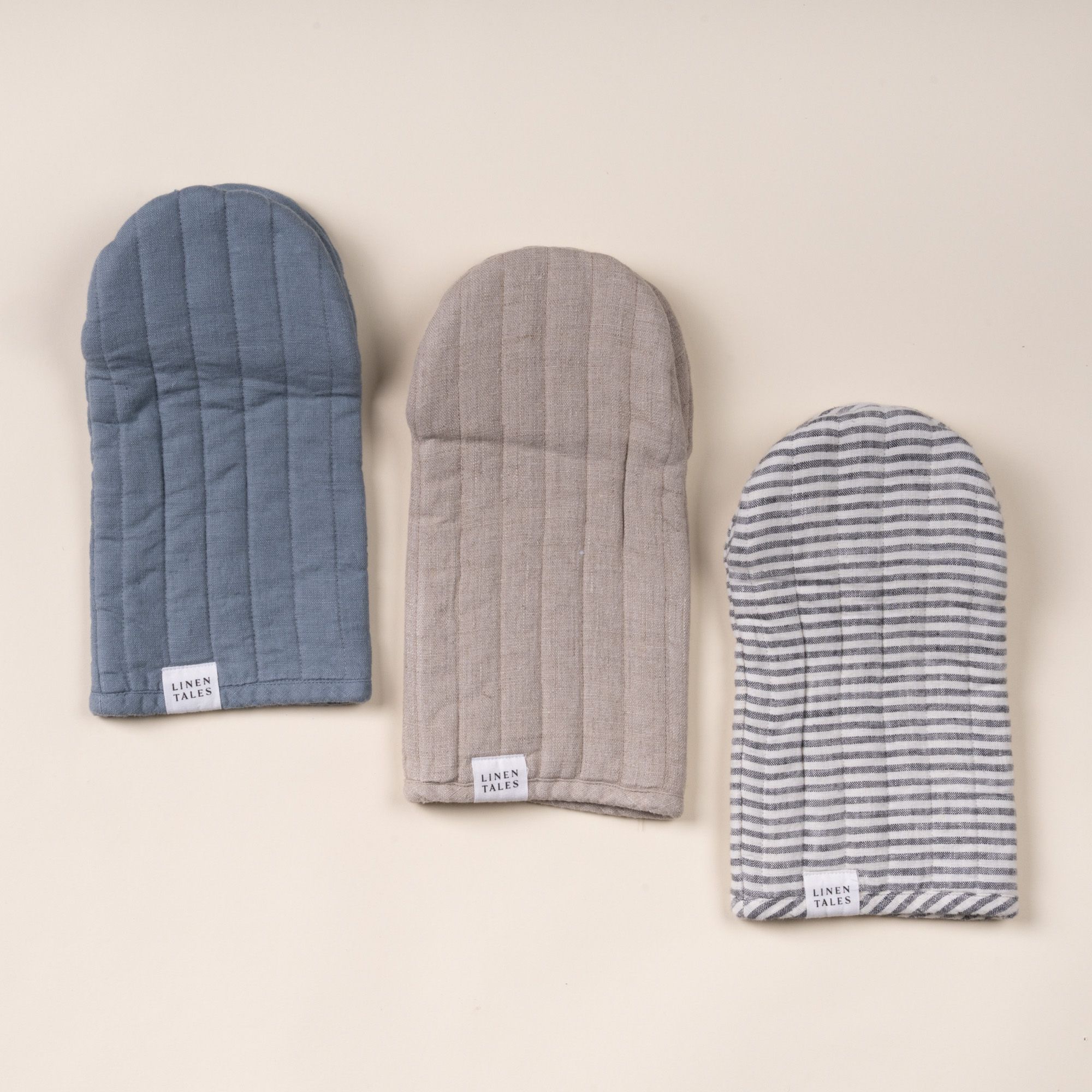 Three Linen Tales oven mitts in blue, beige, and gray-and-white stripes, laid side by side on a light background.